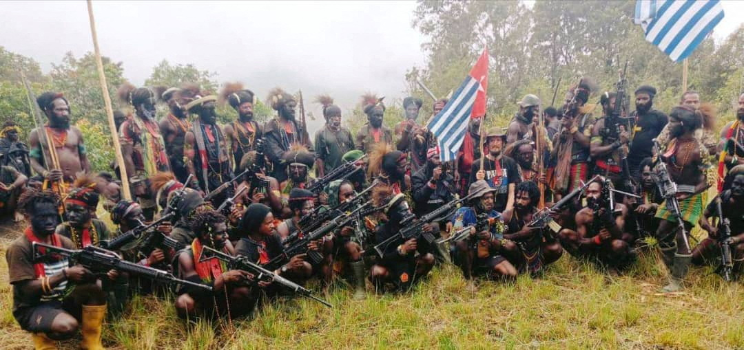 Phillip Mehrtens, a kidnapped NZ pilot, sitting with his captors somewhere in Indonesia's Papua province. The fighters are in black and armed with weapons. Some are in more traditional clothing. Mehrtens is sitting in the middle of the group wearing a black shirt with the words Papua written on it. He is holding the Morning Star flag of the independence movement. He looks thinner than in earlier videos and has a beard.