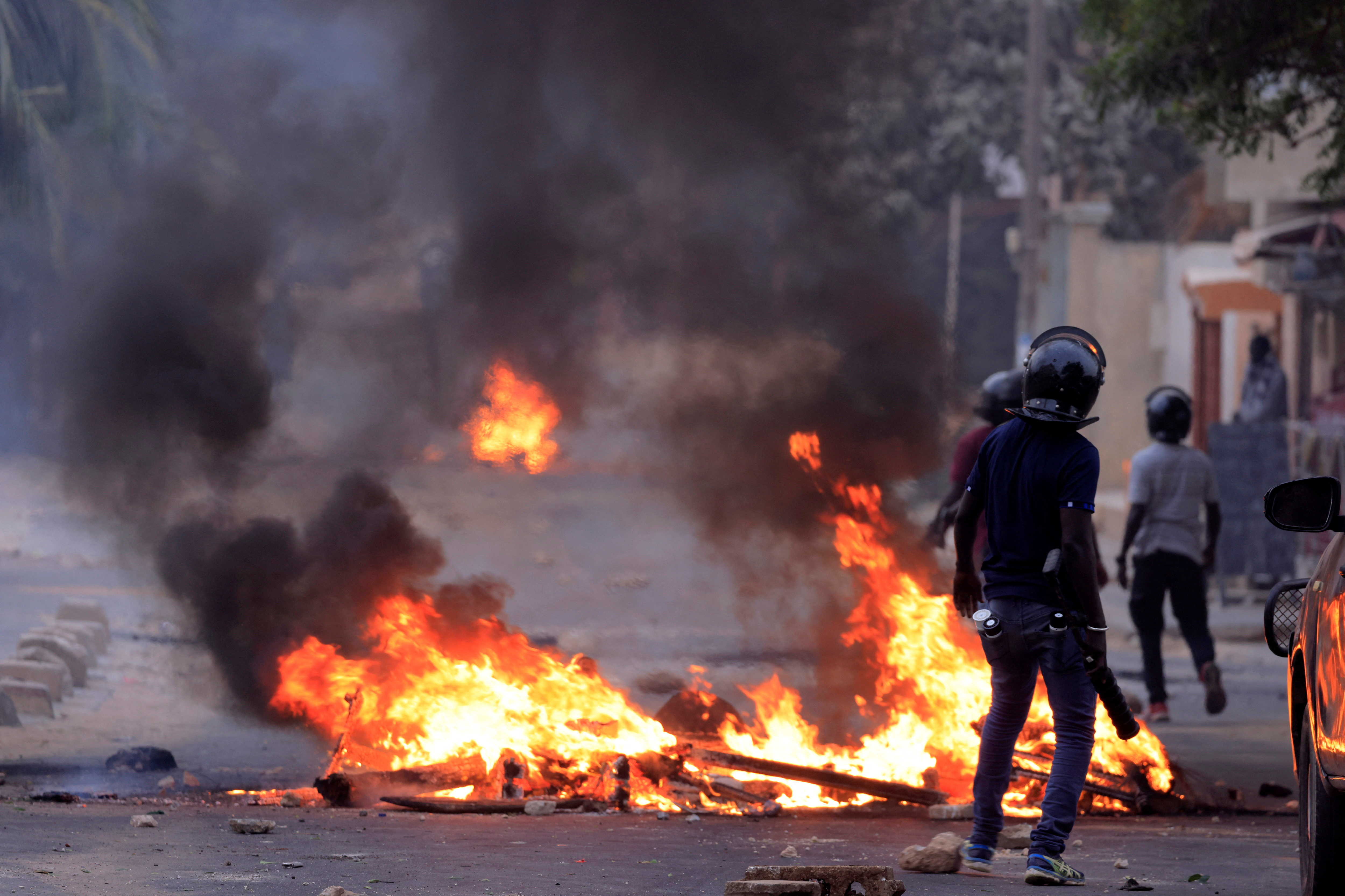 A man in a helmet, dressed in black, passes a burning pile of what appear to be beams, as smoke billows into the sky.