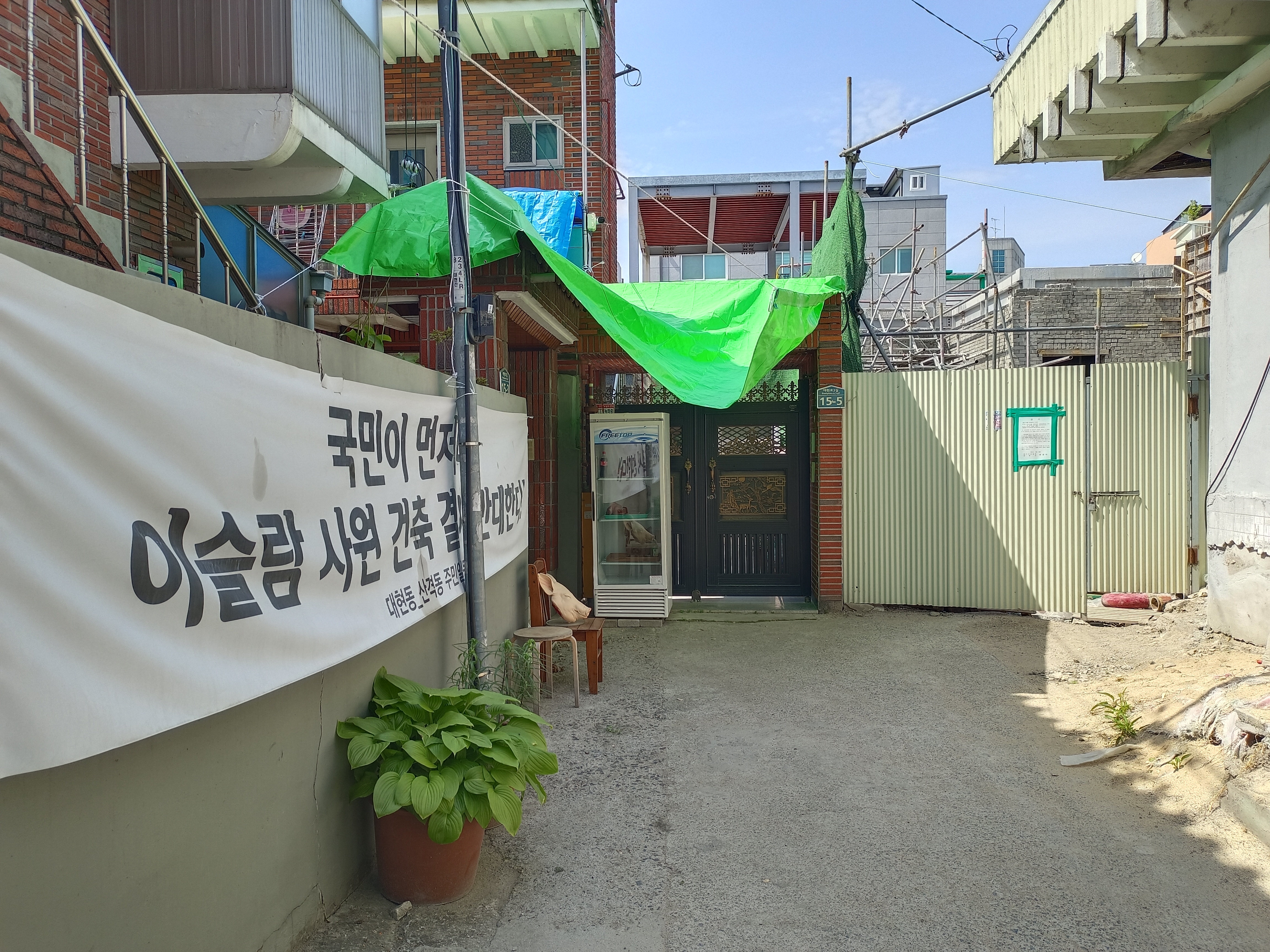 Pathway leading to the mosque construction site. A banner on left reads 'People first! Against the construction of the mosque!' and there is a plastic pig head on a chair in front of it. at the back, next to the site entrance is a refrigerator containing three pig heads. The construction site is on the right. Scaffolding is visible.