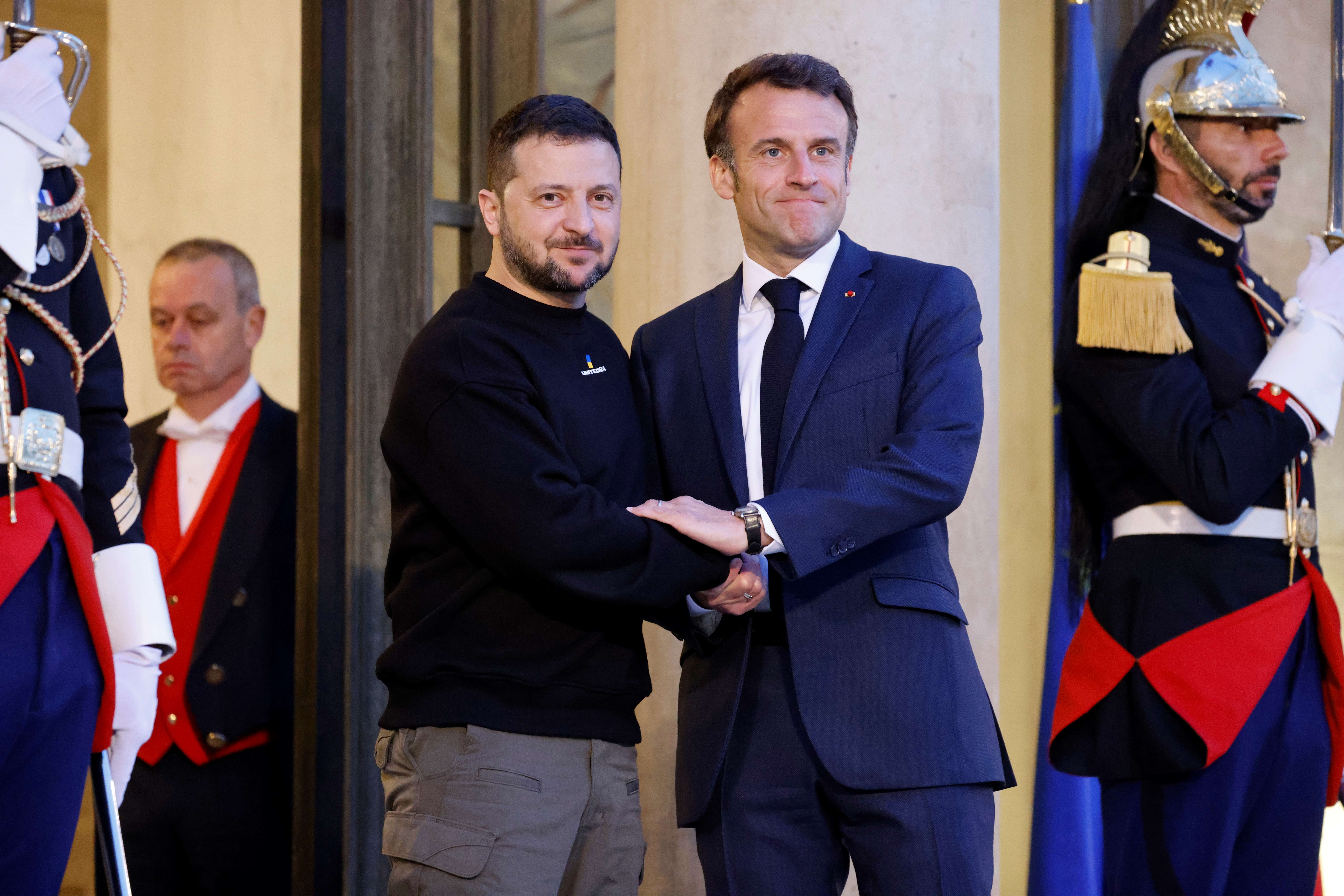French President Emmanuel Macron greets Ukrainian President Volodymyr Zelenskyy at the Elysee Palace. Macron is in a blue suit and tie, while Zelenksyy is in a black sweath short and khaki trousers. They look resolute.