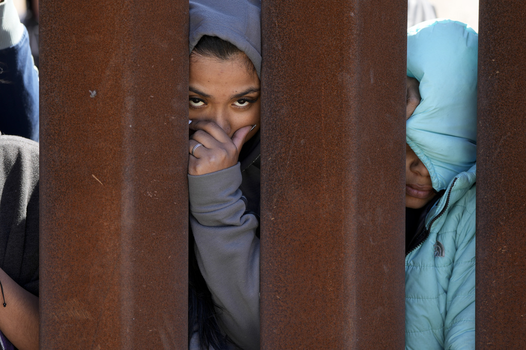 Migrants waiting to apply for asylum between two border walls look through the wall