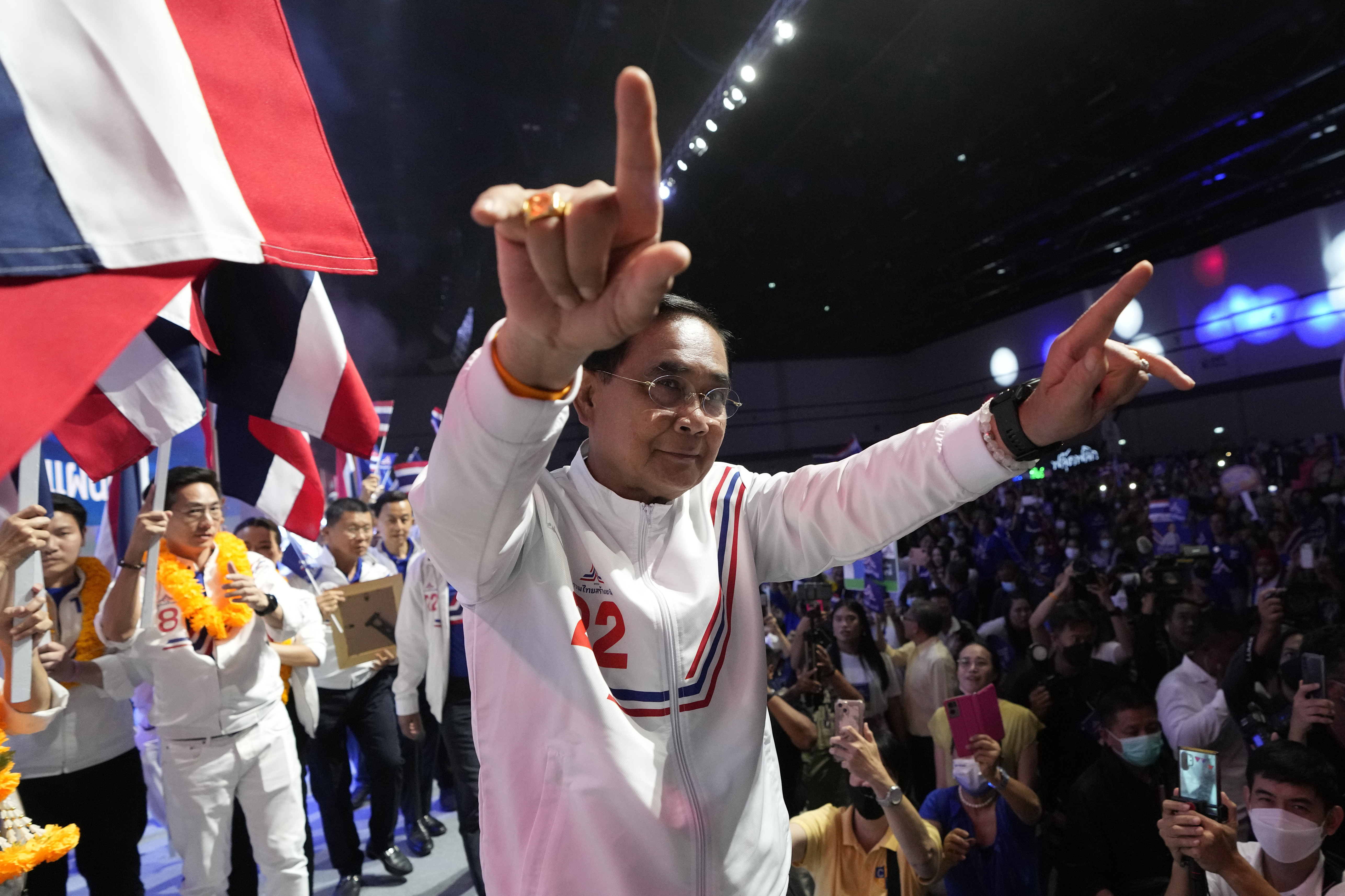 Prayuth gesturing to his supporters. He is wearing a track suit in the party colours of red white and blue and there are flags in the background.