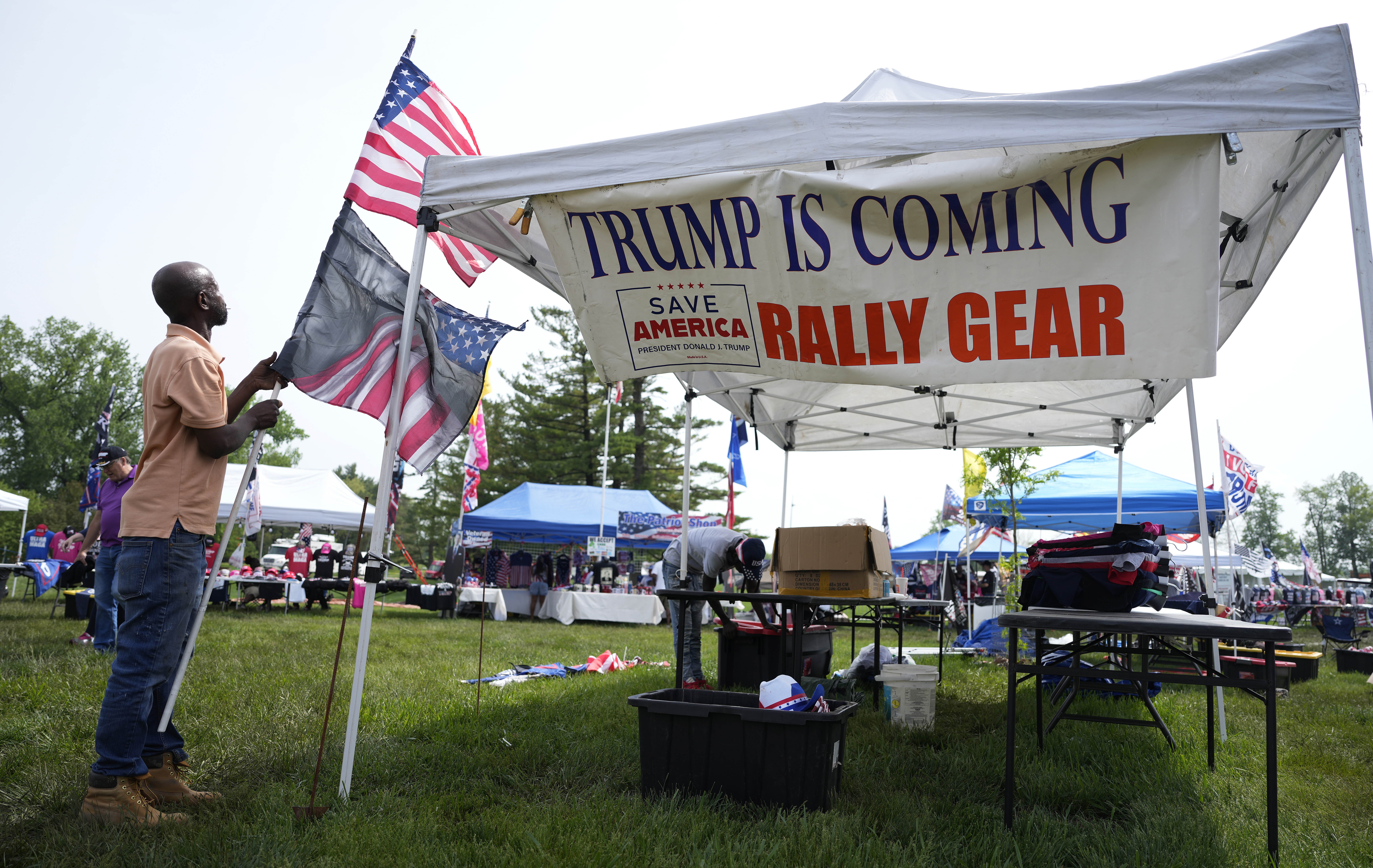 A man disassembles a stack of flags, next to a white tent with a banner that reads: "Trump is coming. Rally gear."