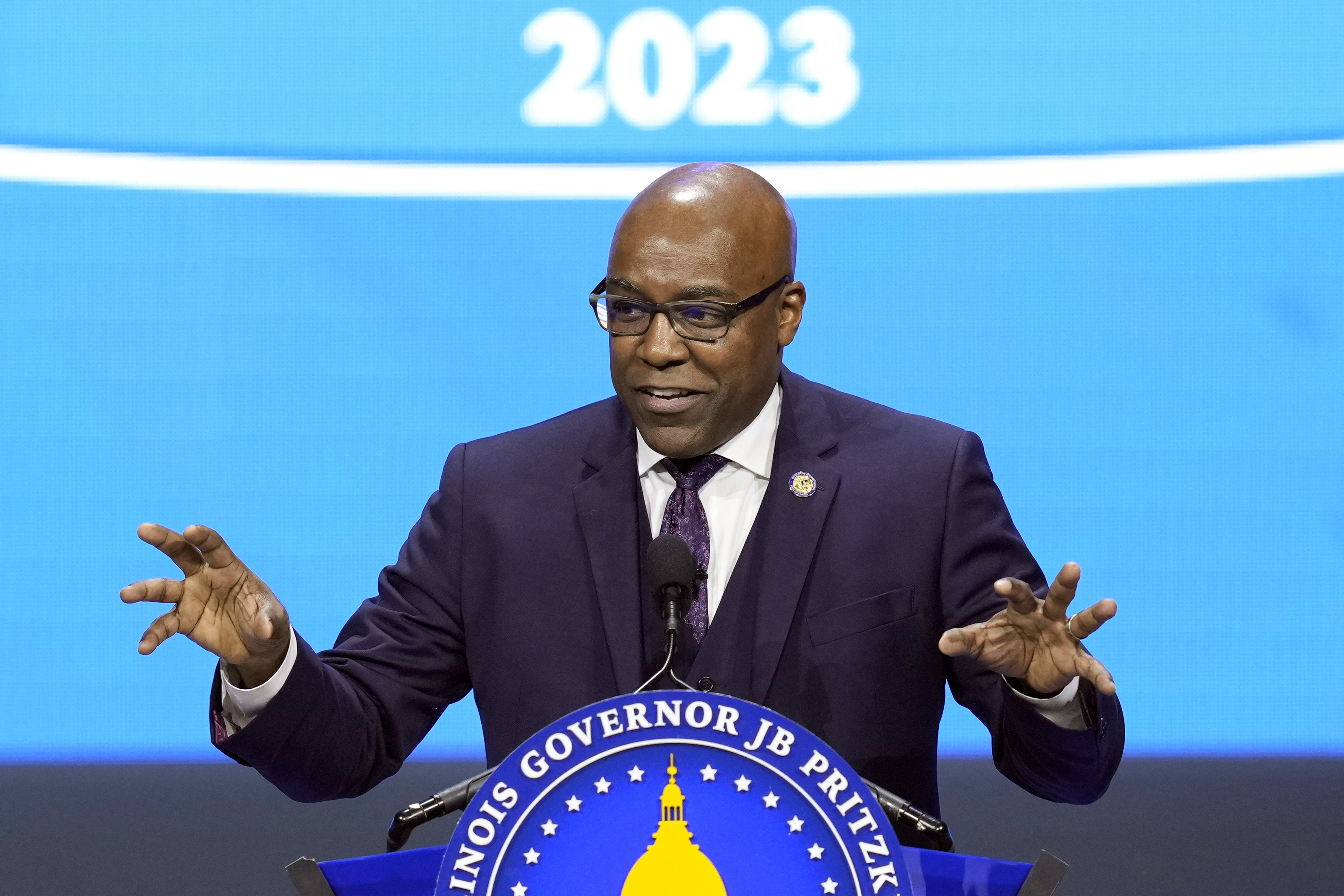Kwame Raoul, wearing a blue suit, gestures with both hands behind a podium. He is standing in front of a backdrop that reads "2023."