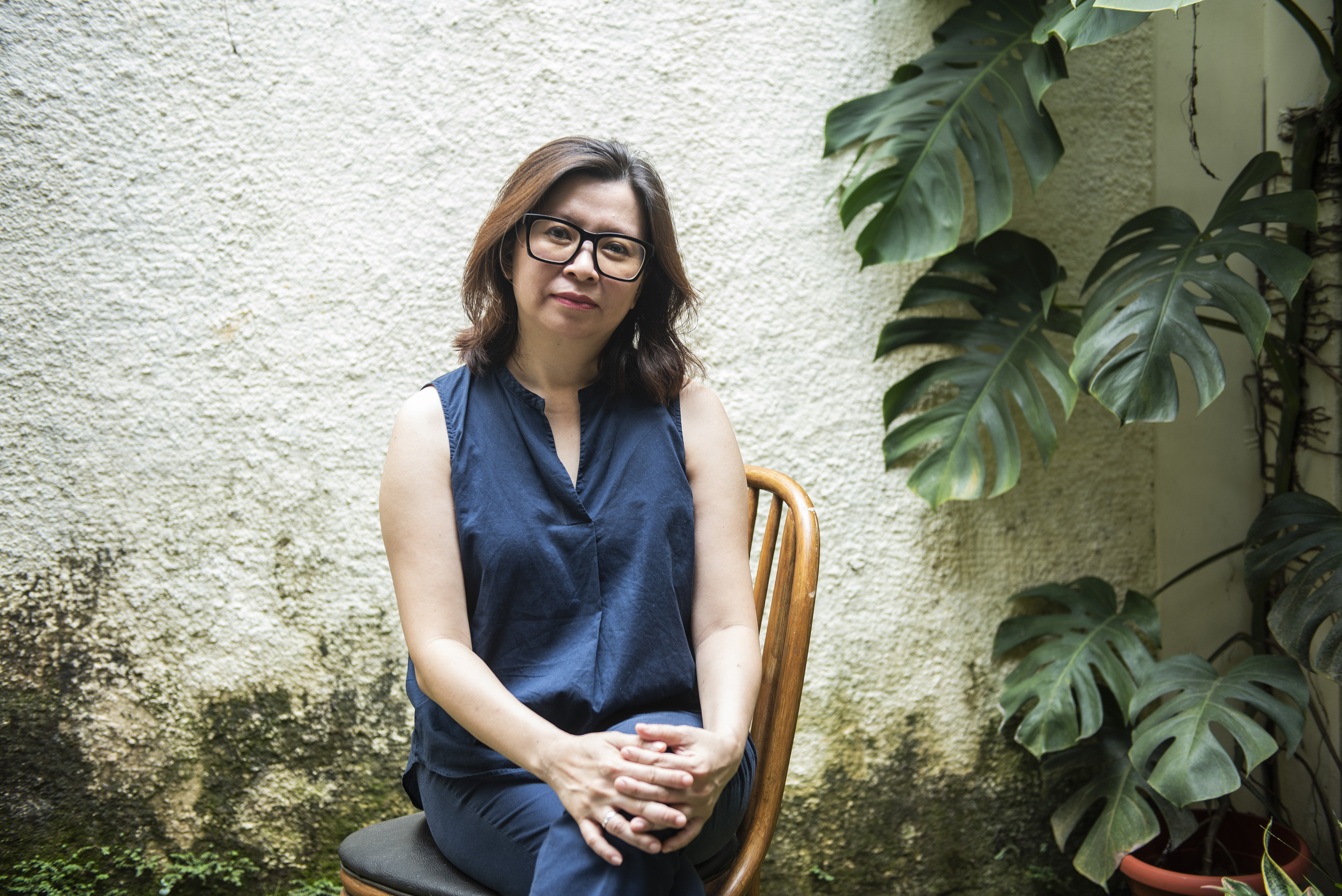 Evi Mariani pictured sitting in a chair. Sh eis wearing a blue dress and has her hands clasped around her knees. She looks happy. There's a large cheese plant nearby.