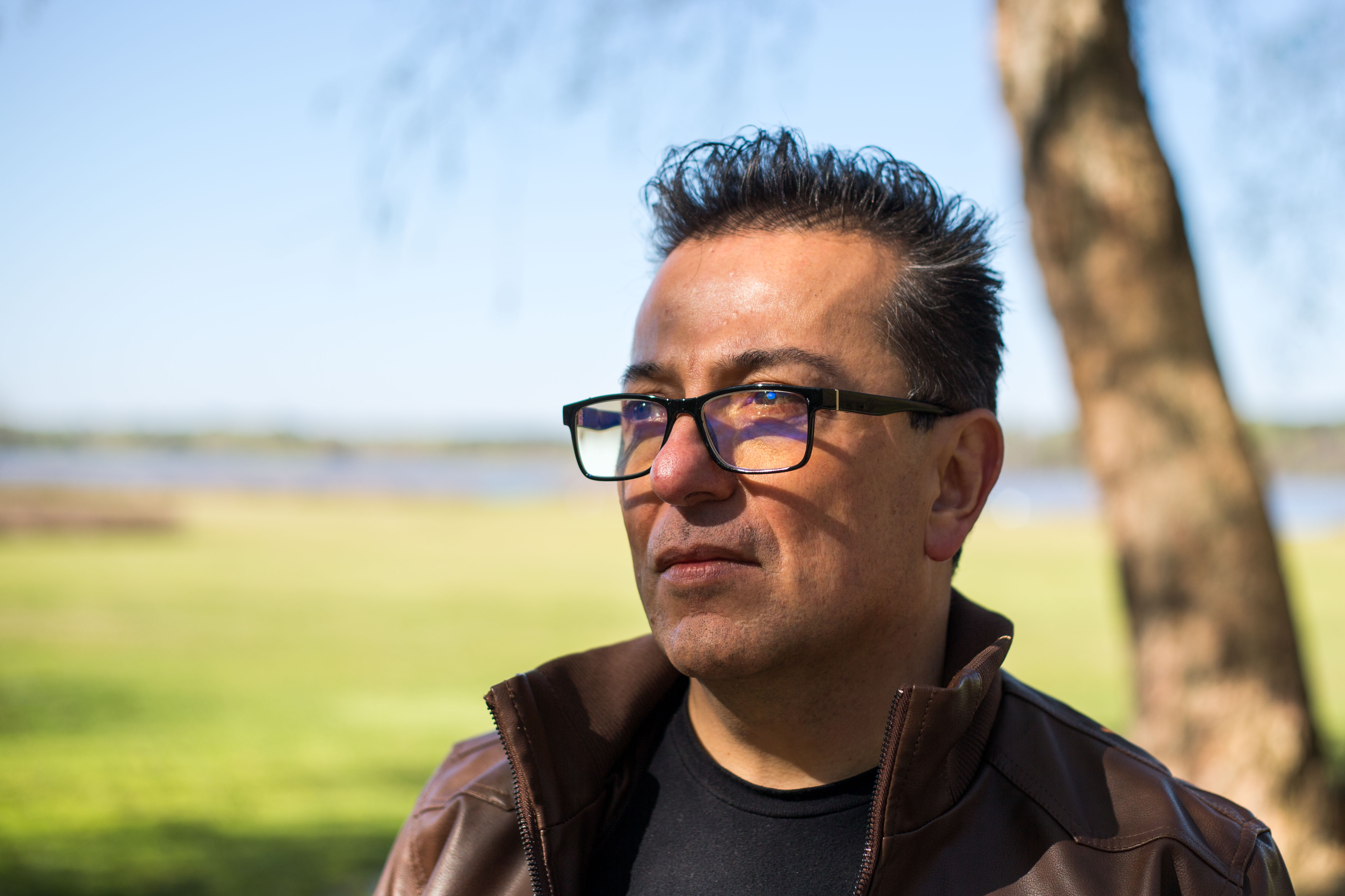A close-up of a man in glasses and a brown pullover sweater, as he stands outside.