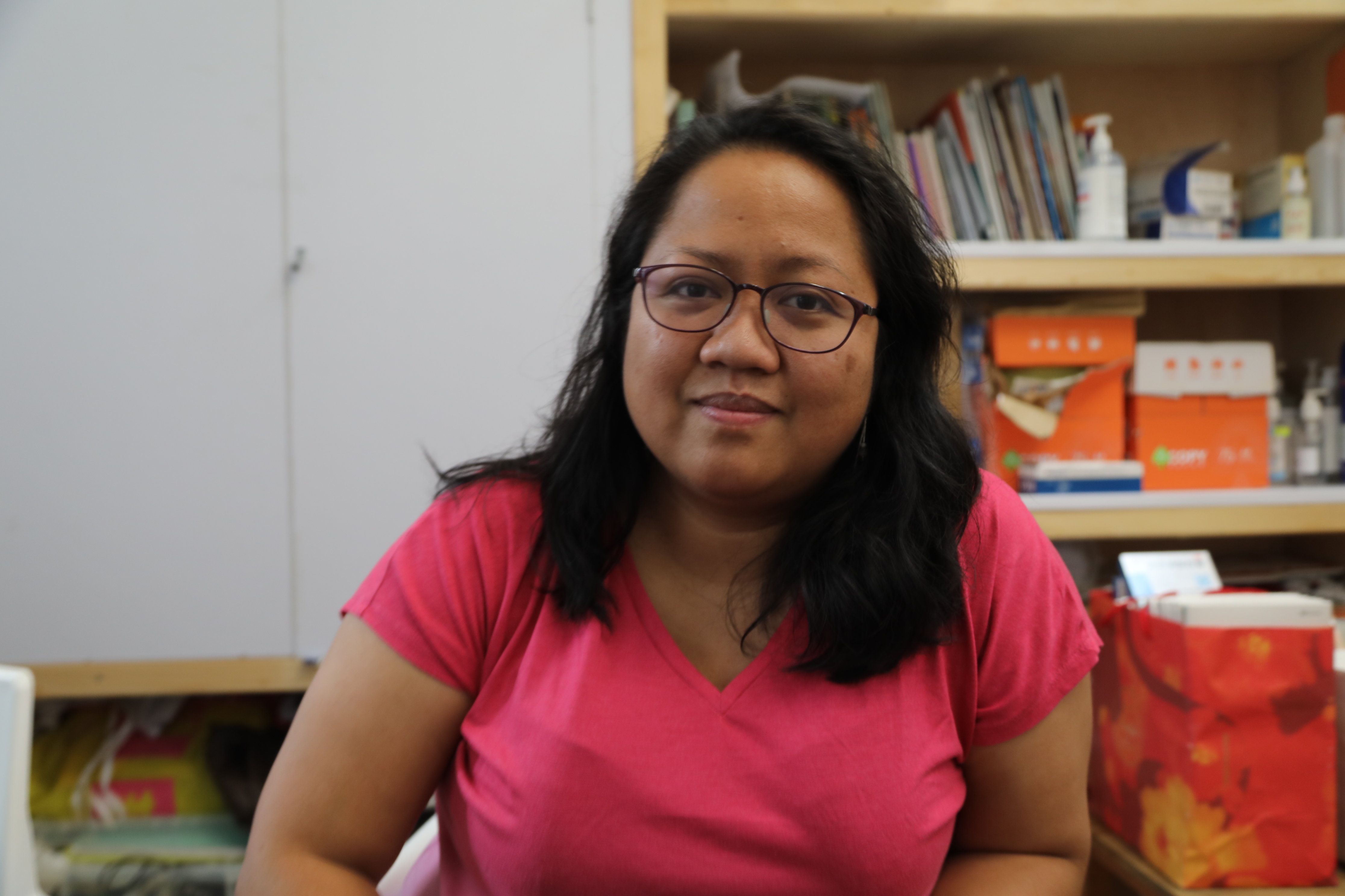 A portrait of Shiela Tebia-Bonifacio. She is sitting at a desk with a bookcase behind her.