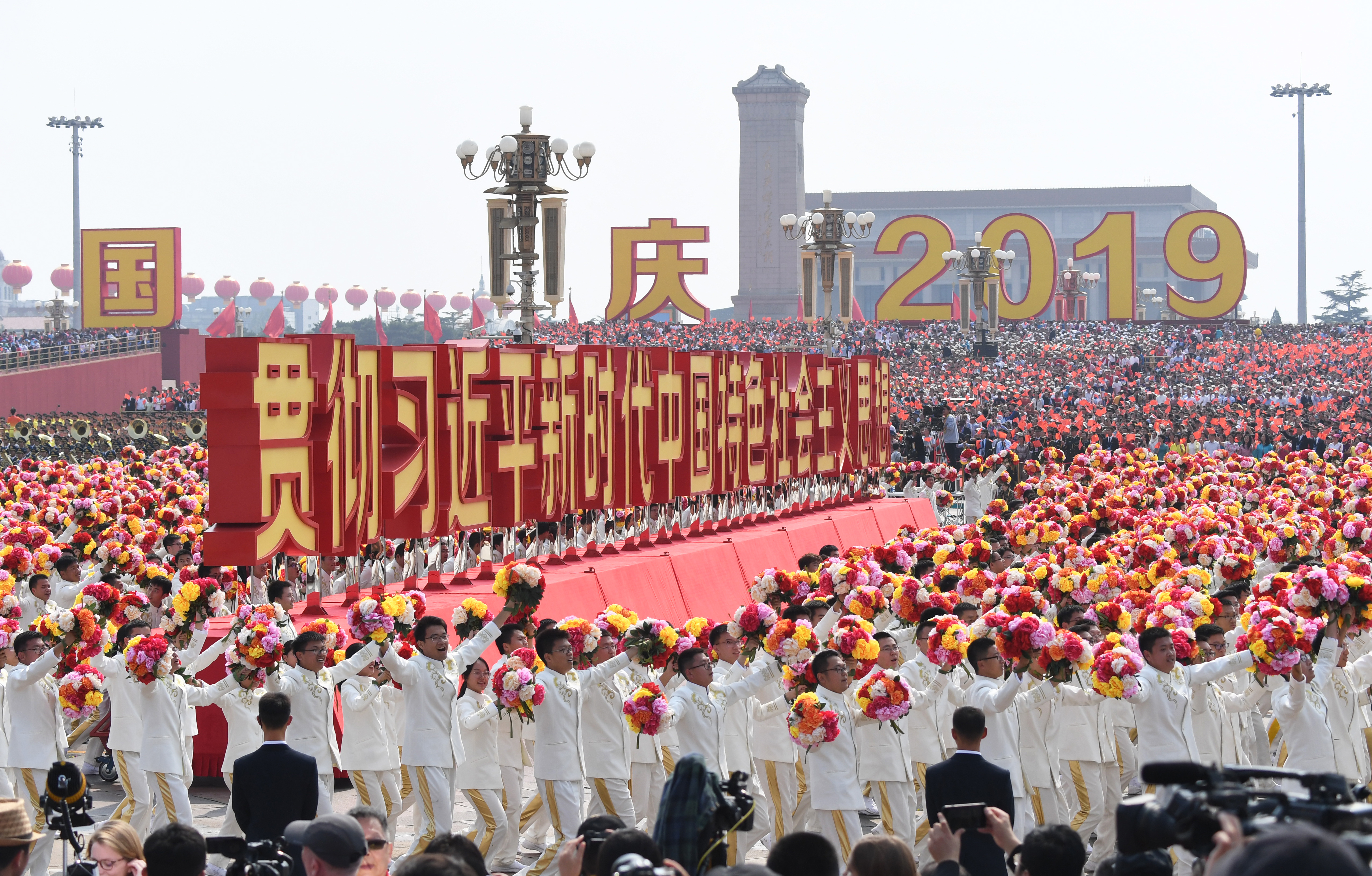 A large float spelling out "Implement Xi Jinping Thought on Socialism with Chinese Characteristics for a New Era" in huge red Chinese characters passes through Tiananmen Square during the 2019 parade to mark the 70th anniversary of the founding of the People's Republic of China. There are people dressed in white and carrying red, yellow and pink flowers walking in front of it