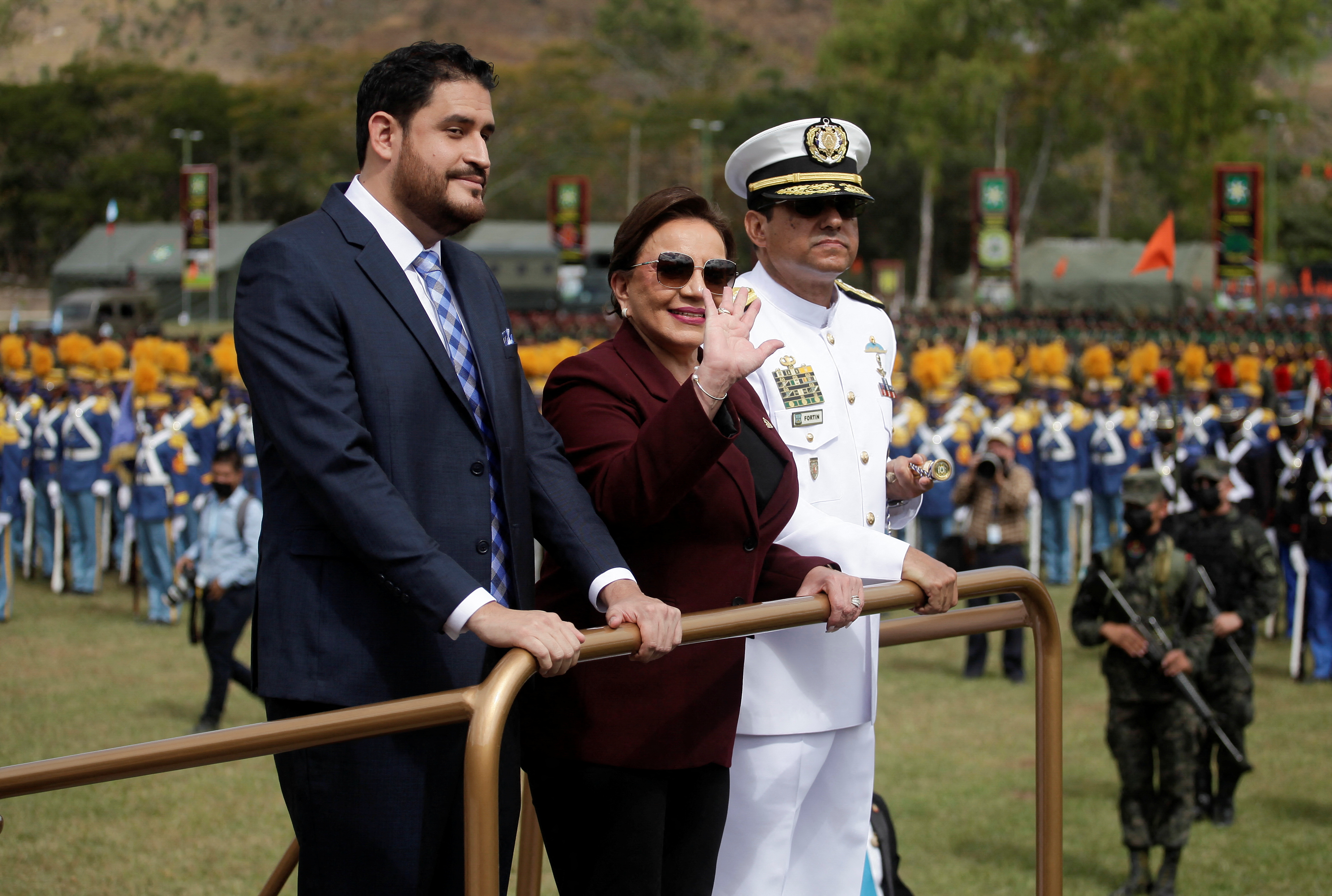 Three officials pose by a railing: one in a suit and tie, another in a pants suit and the third in a white military dress uniform.