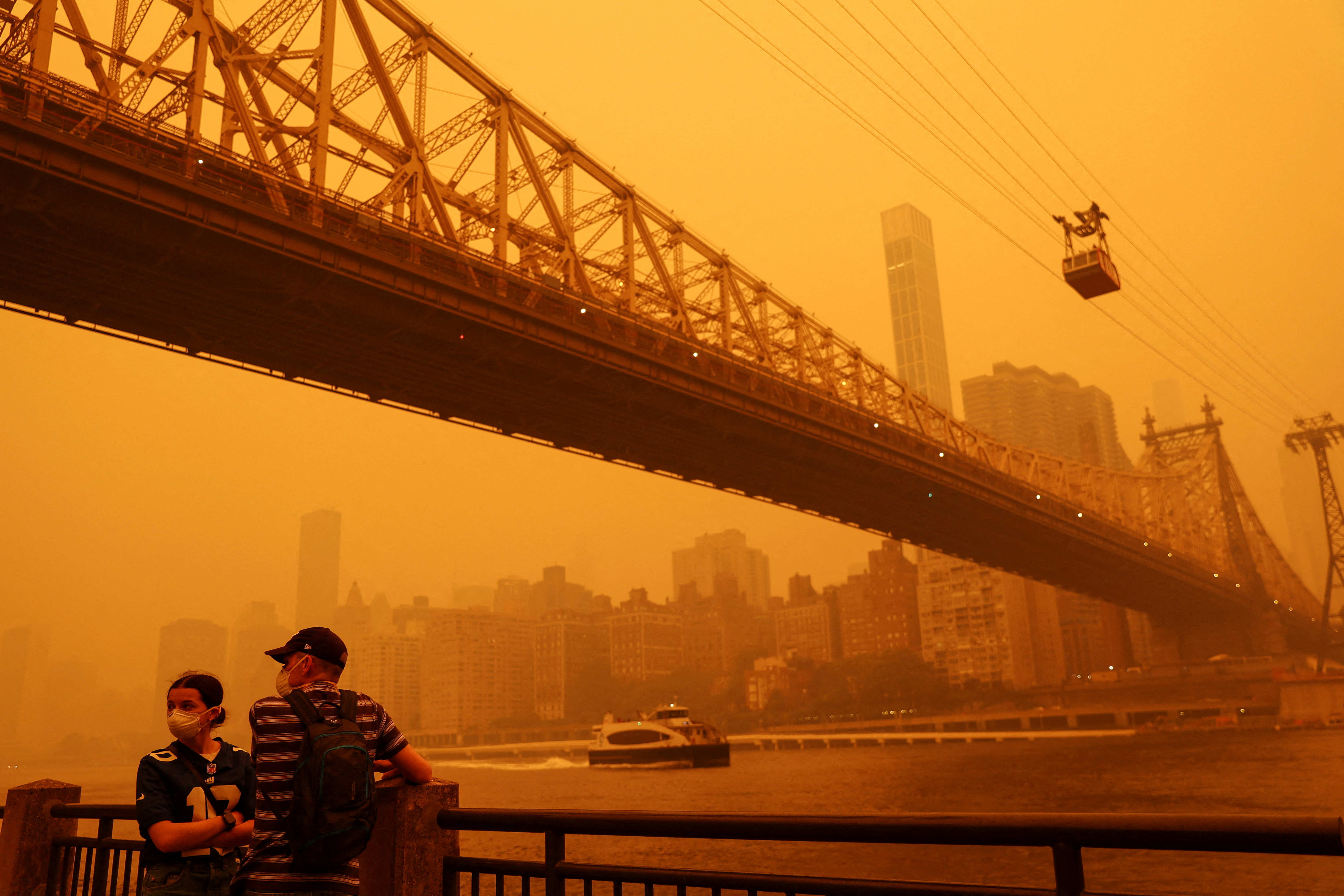 People wear protective masks as the Roosevelt Island Tram crosses the East River while haze and smoke from the Canadian wildfires shroud the Manhattan skyline in the Queens Borough New York City