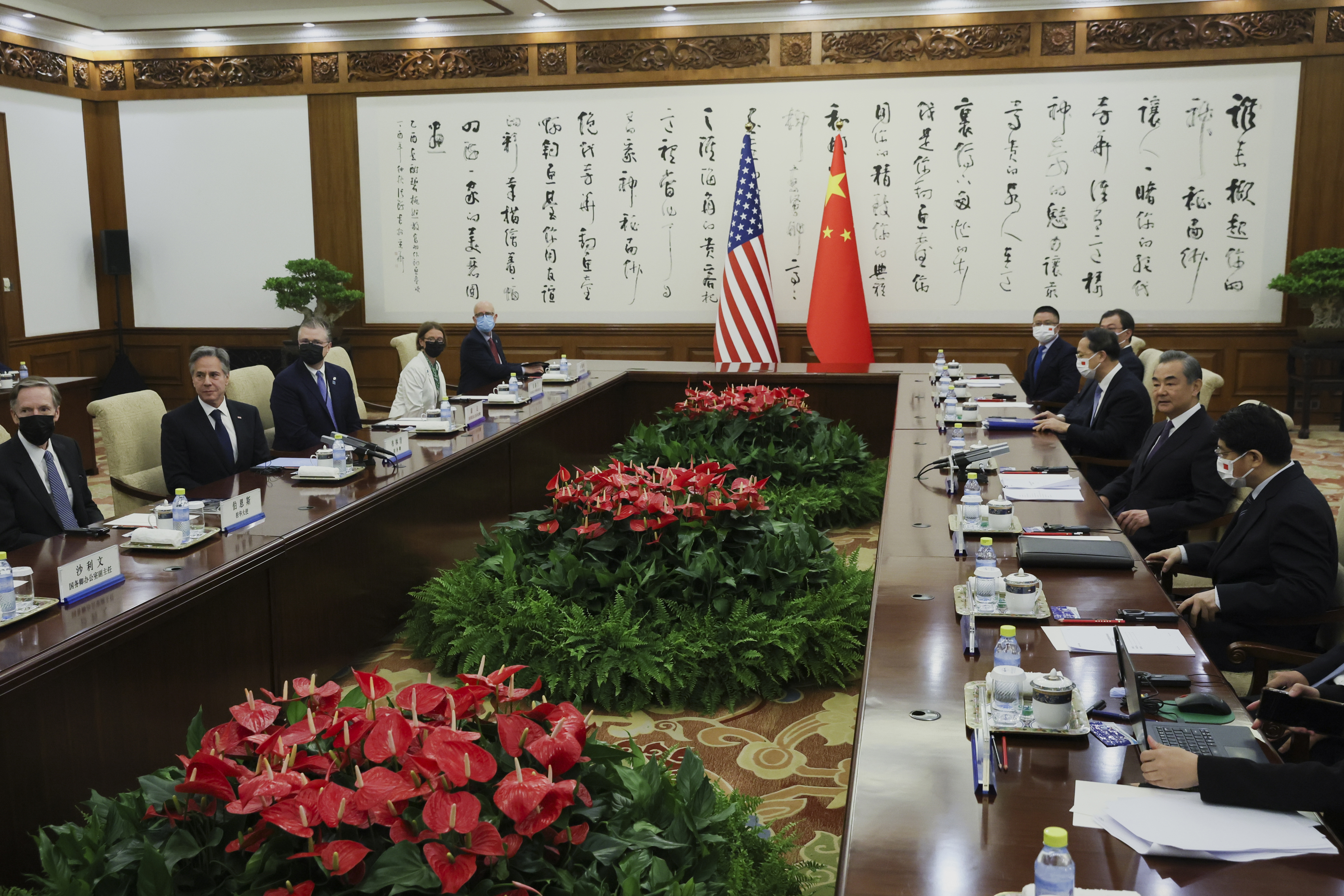 The US and Chinese delegations at the table. They are sitting opposite each other on long tables. There are plants in the middle. The countries flags and Chinese calligraphy are behind.