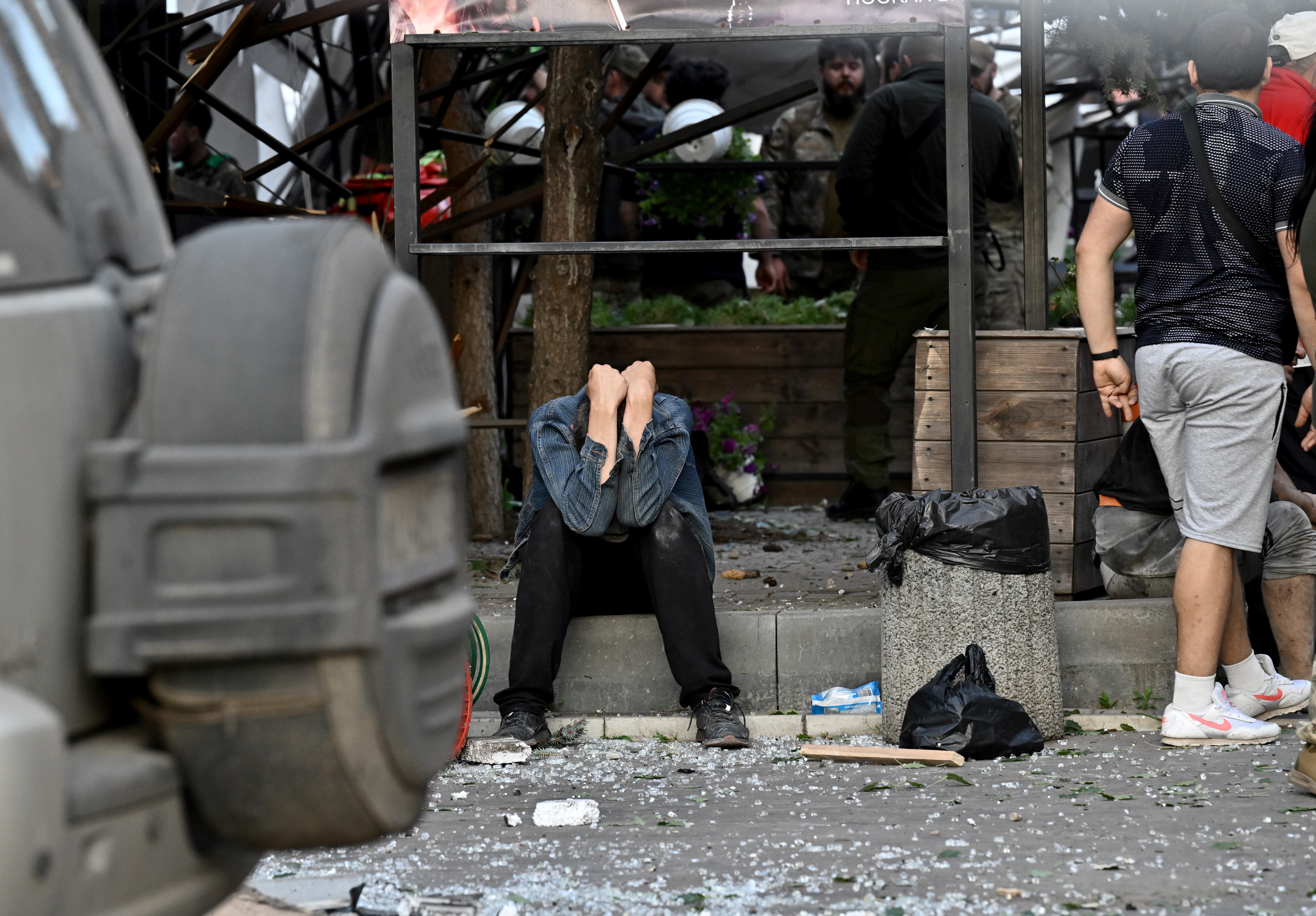 A man covers his face with his hands outside the destroyed restaurant. He is sitting on the side of the road. There is debris around and in front of him. He looks in despair.