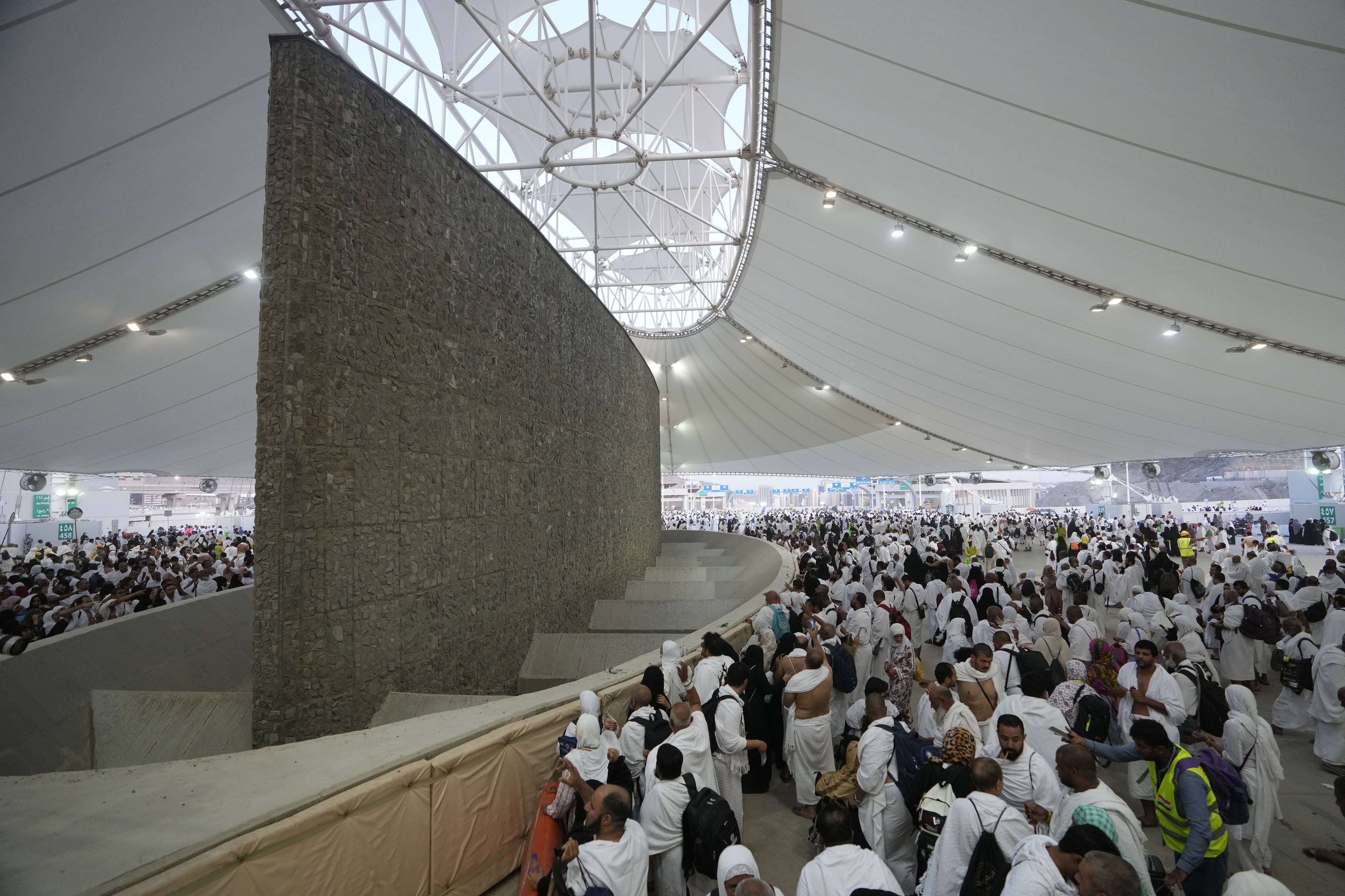 Pilgrims dressed in white gather under a sunshade to throw rocks at a big concrete pillar, one of the jamarat
