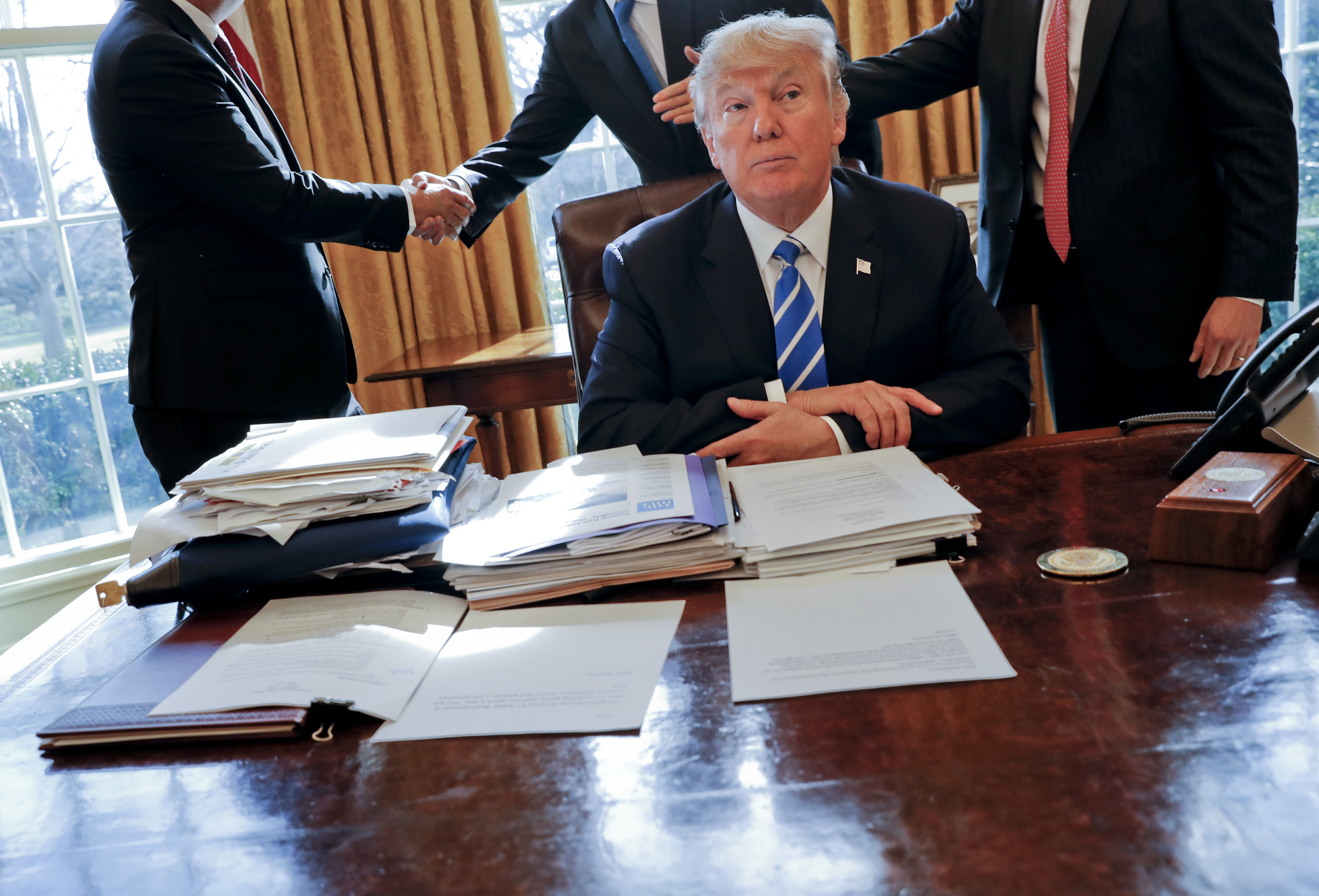 Donald Trump at the Oval Office desk when he was president. There is a pile of documents in front of him and to his right. People are shaking hands behind him.