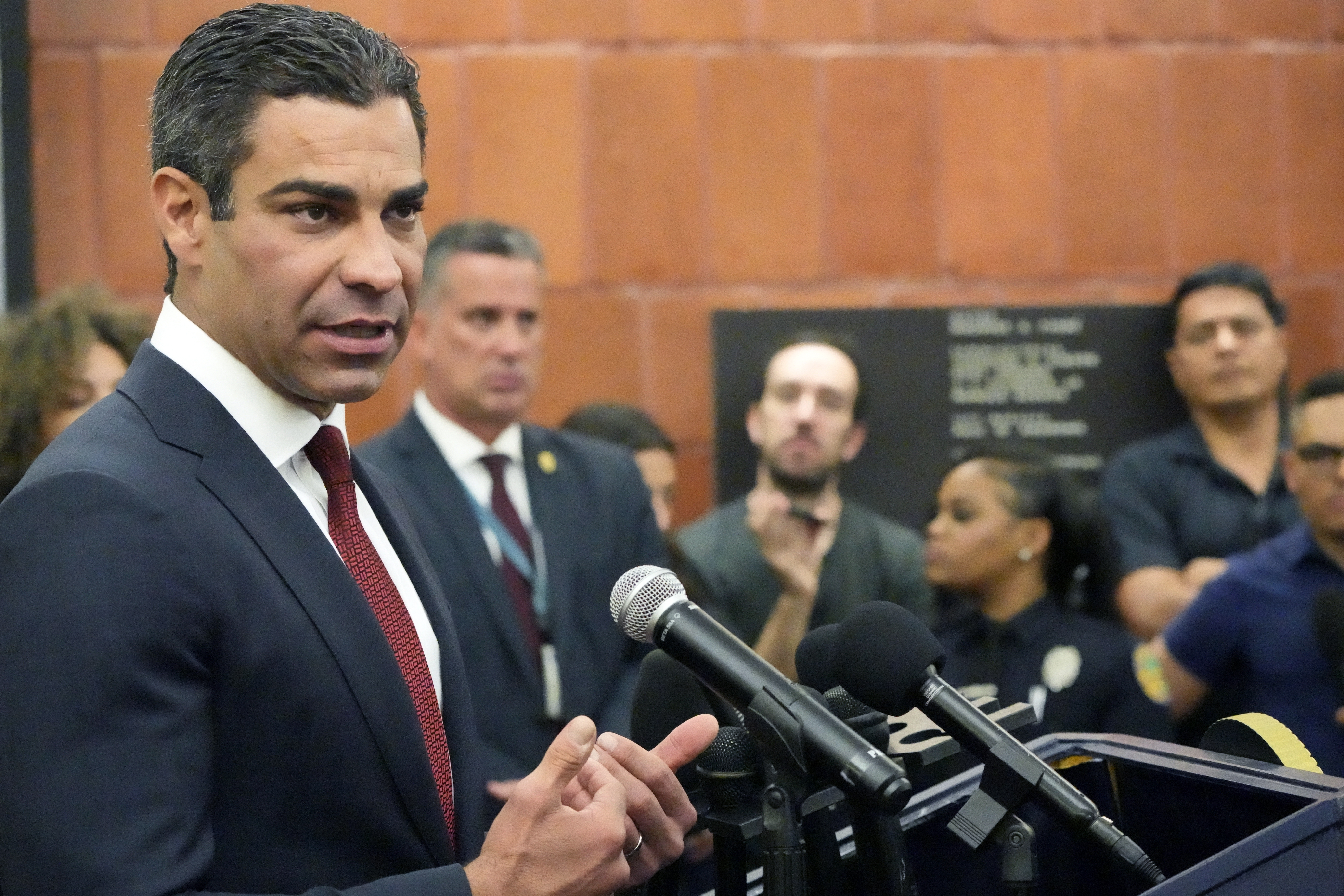 A man stands in a wood-paneled room at a podium, speaking into a microphone as others look on.
