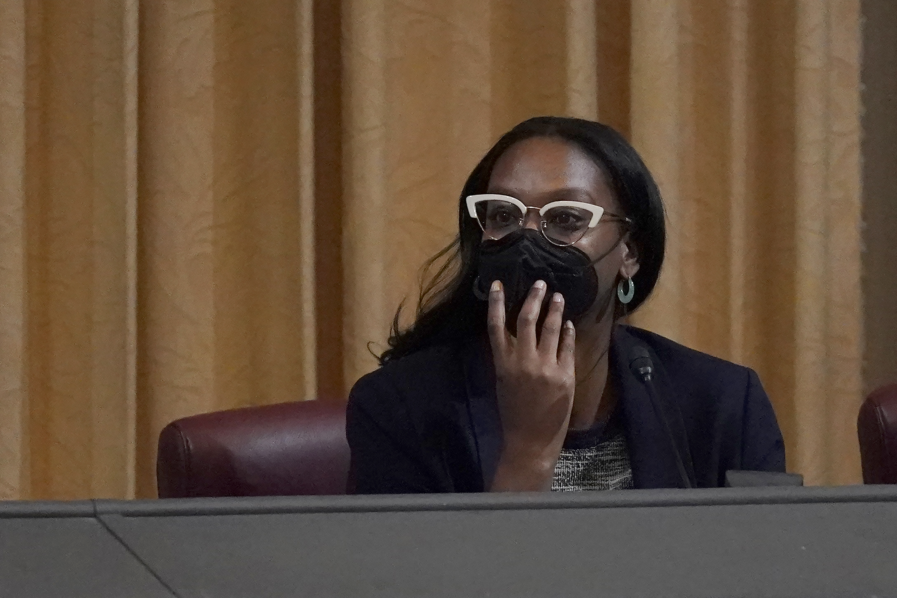 A woman, wearing a face mask and glasses, leans across a panel table, listening.