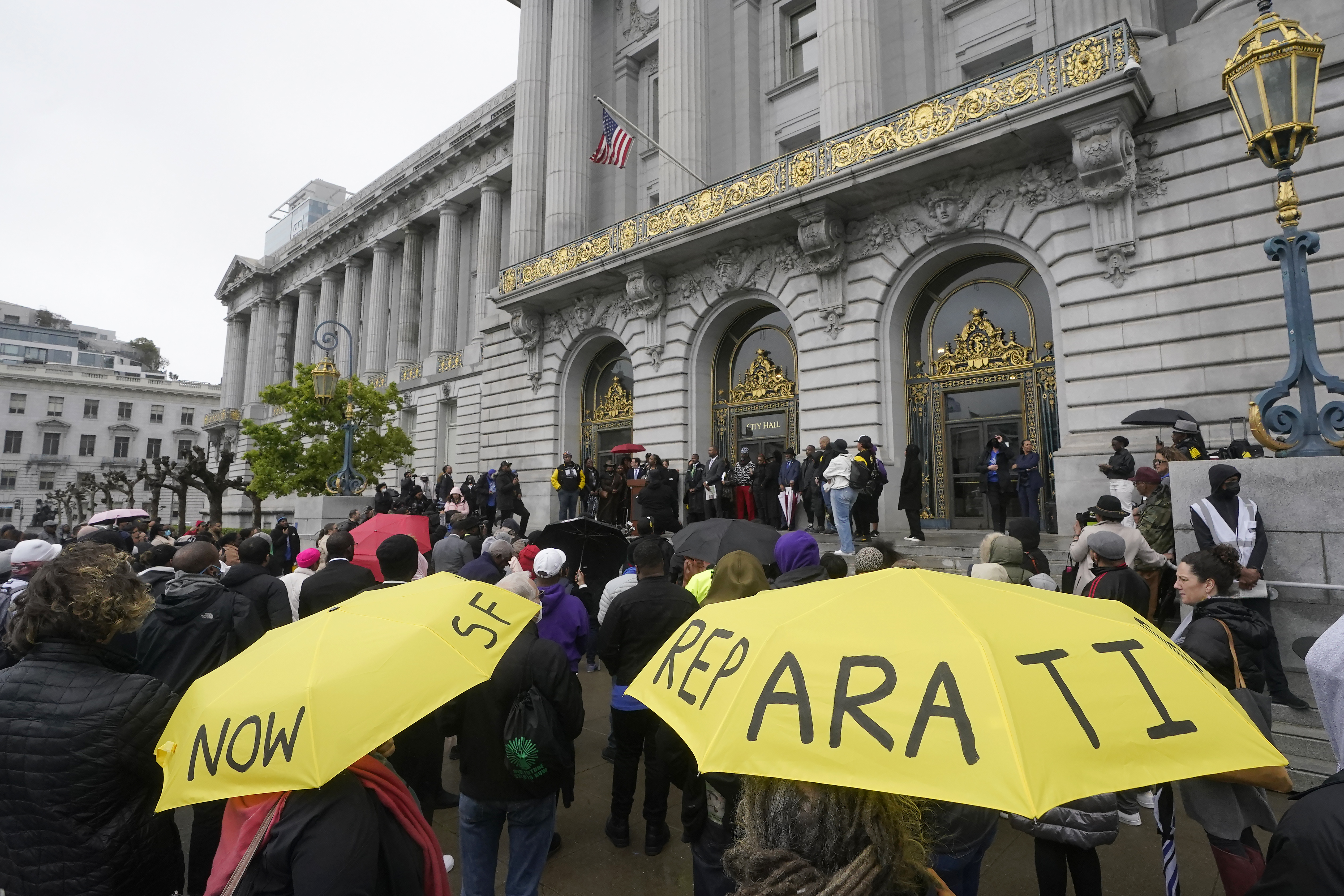 Outside San Francisco's city hall, a crowd has gathered. Two people hold yellow umbrellas, with the handwritten words "reparations now" inscribed on them.