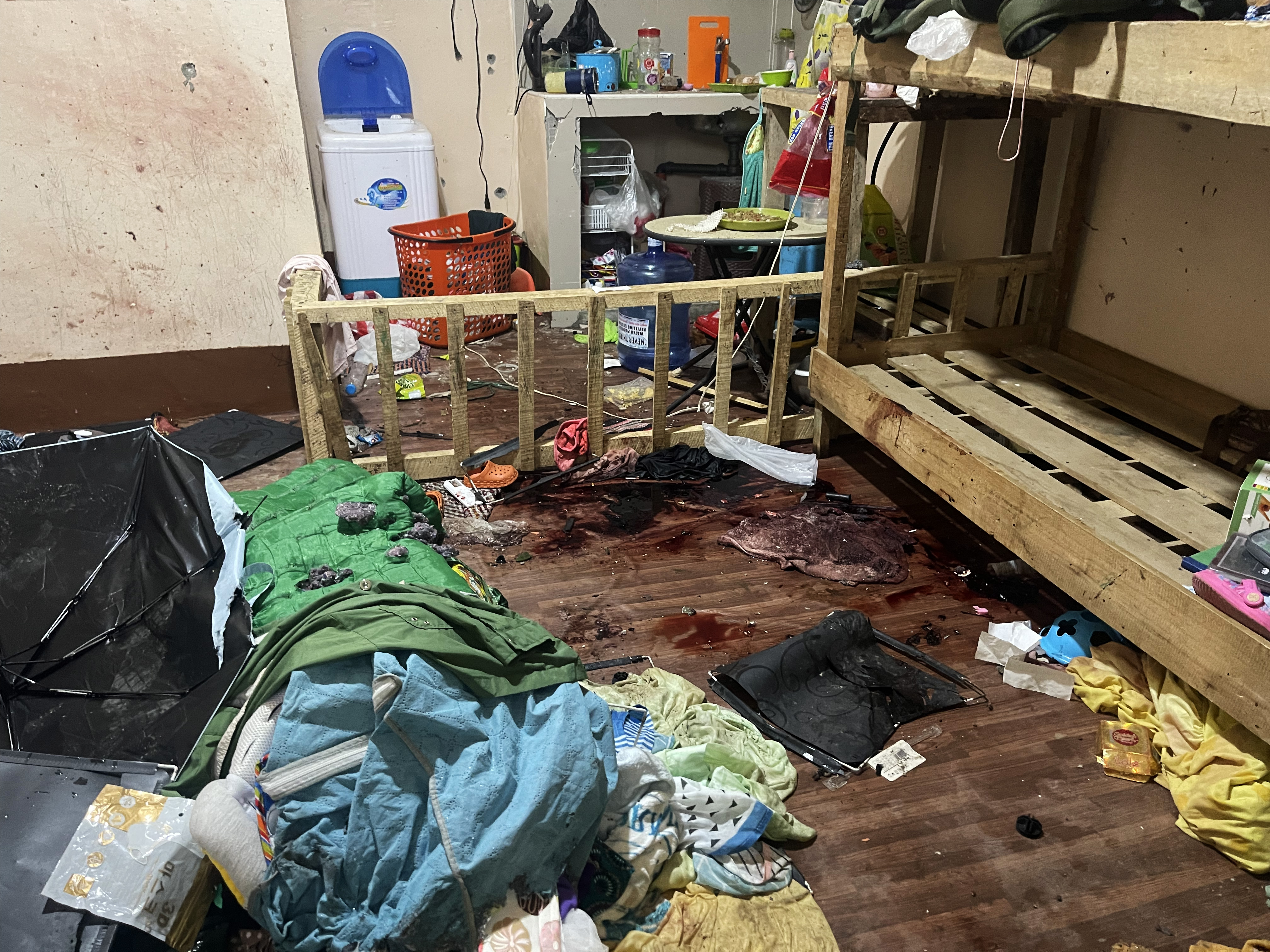 Blankets, sleeping bags strewn about after a raid on the hideout of Abu Zacharia. There are blood stains on the wooden cot. There is a cooking area and water cooler behind.