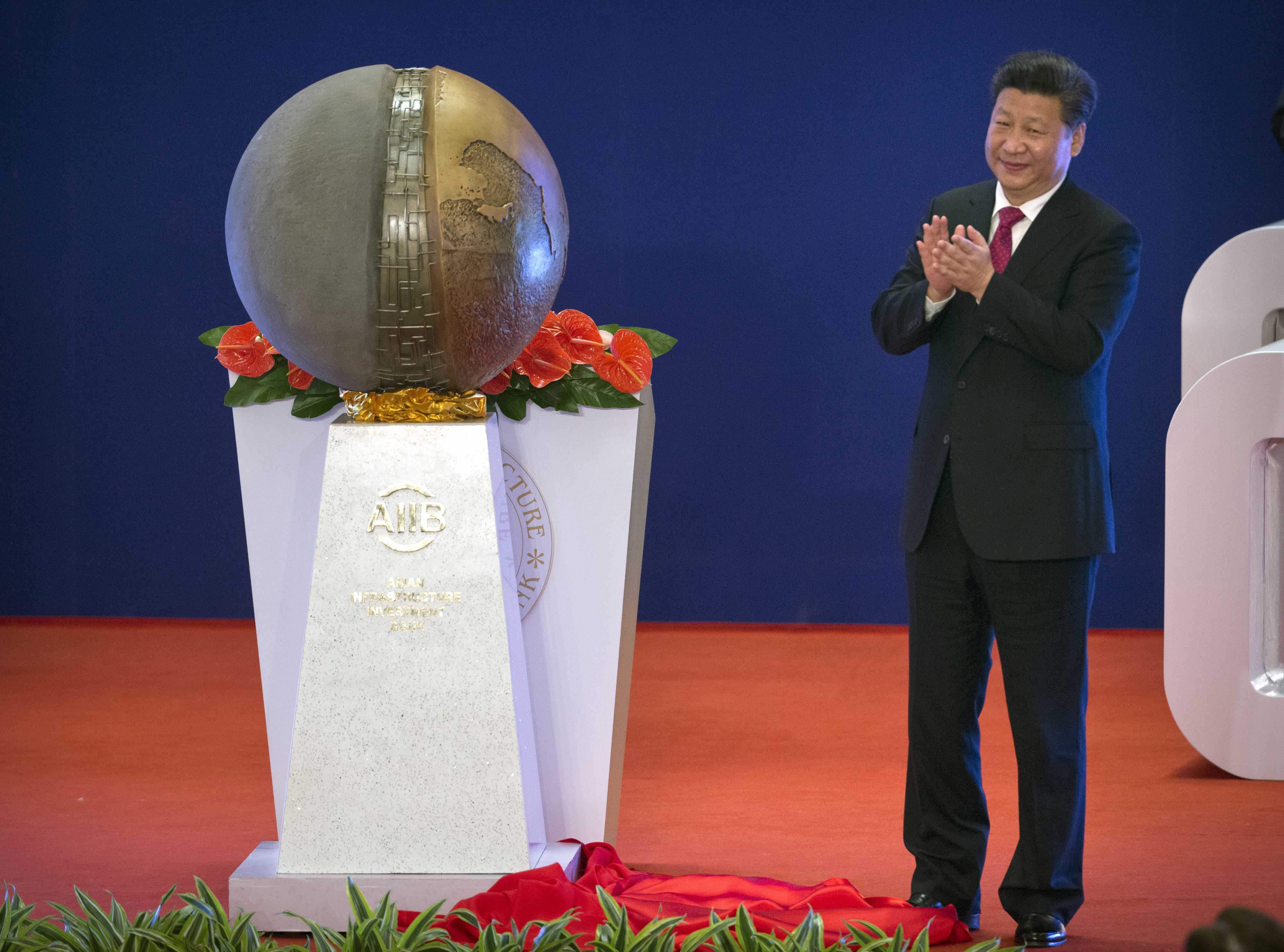 Xi Jinping clapping and smiling at the official launcn of the AIIB. There is a globe sculpture next to him on a white column with the AIIB's logo.
