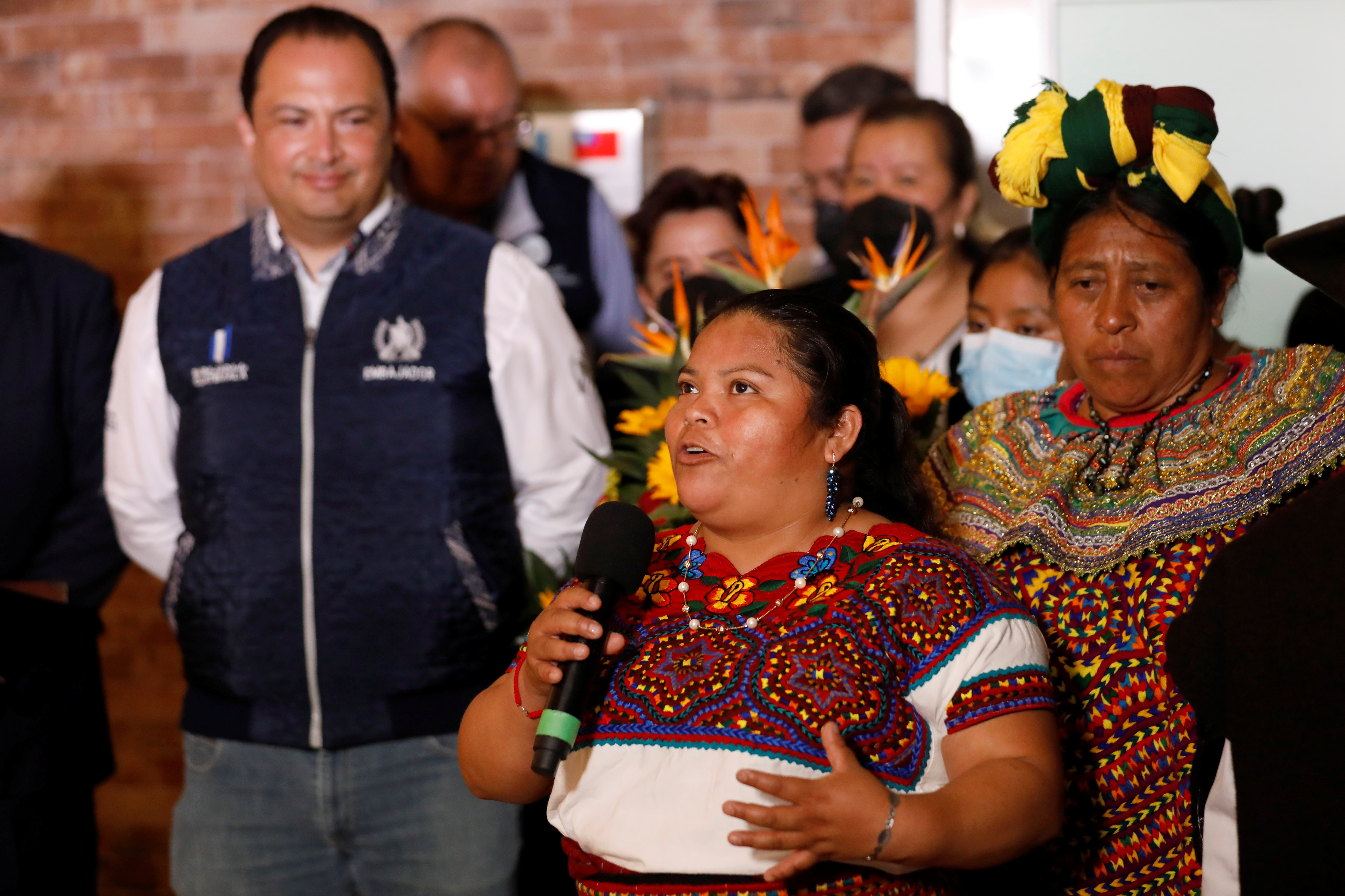 An Indigenous woman in an embroidered top speaks into a microphone as others look on