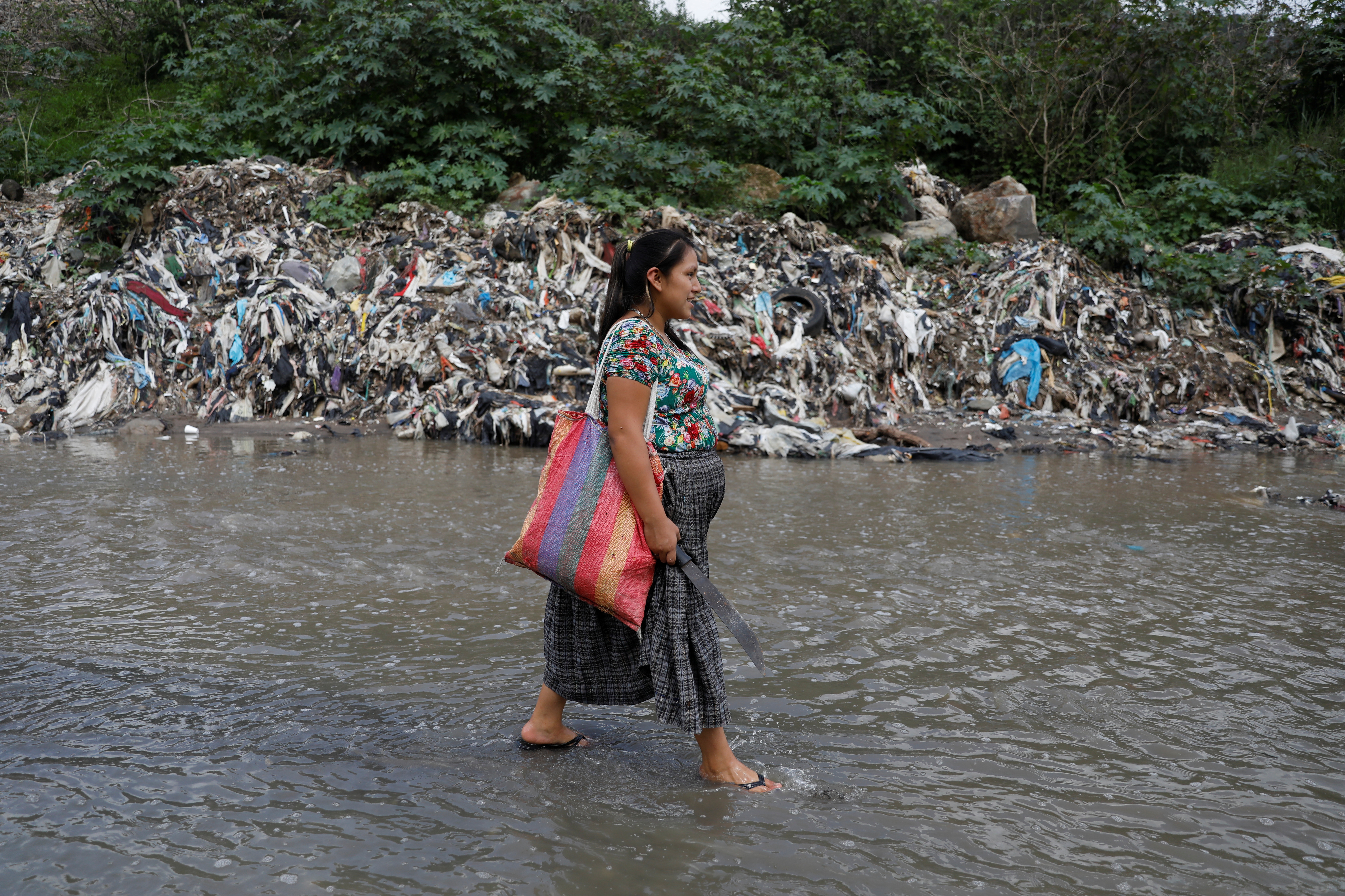A woman with a colorful tote bag walks through the middle of a river, its banks lined with garbage