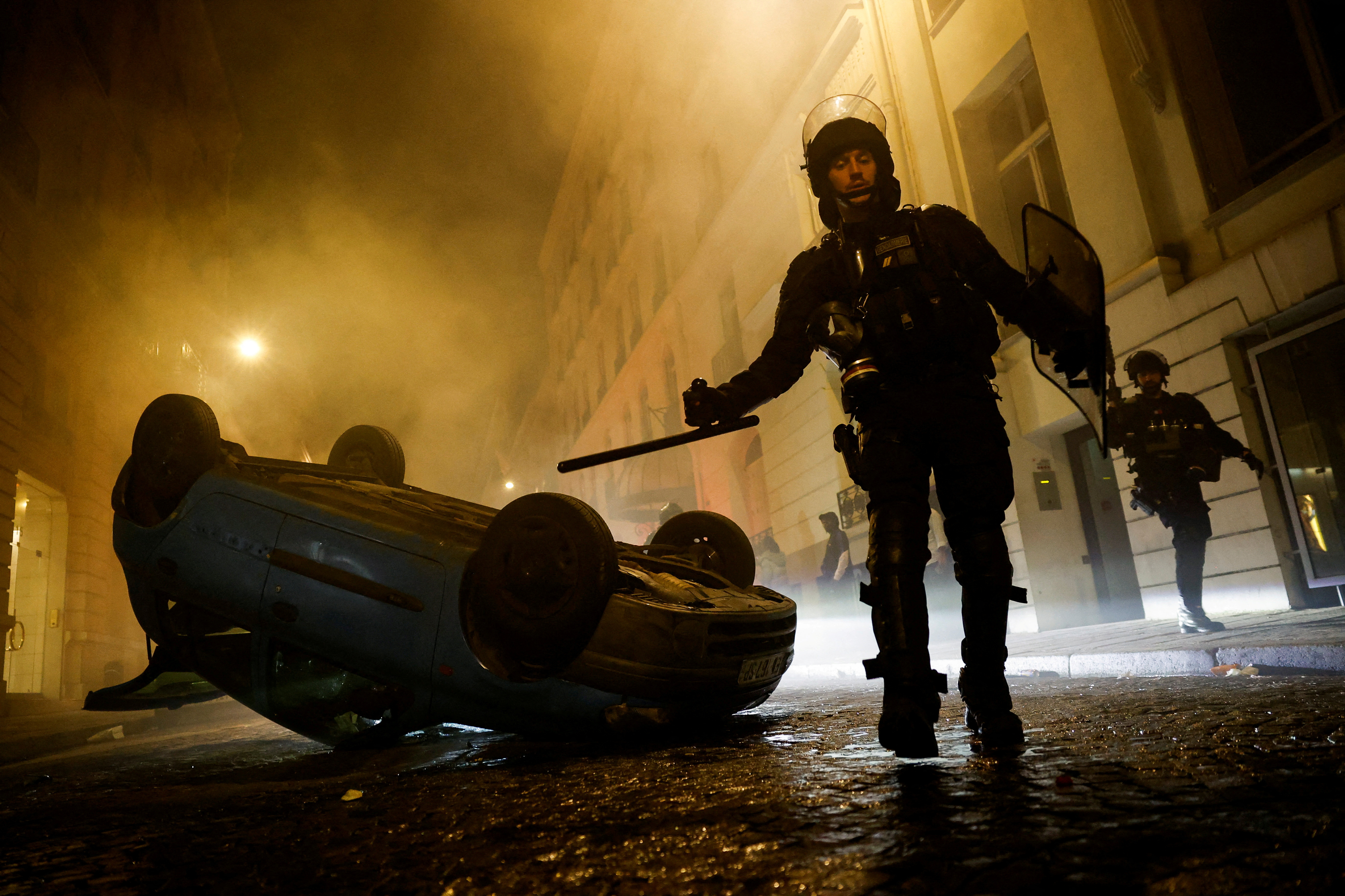 French riot police officers walk next to a vehicle upside down during the fifth day of protests following the death of Nahel, a 17-year-old teenager killed by a French police officer