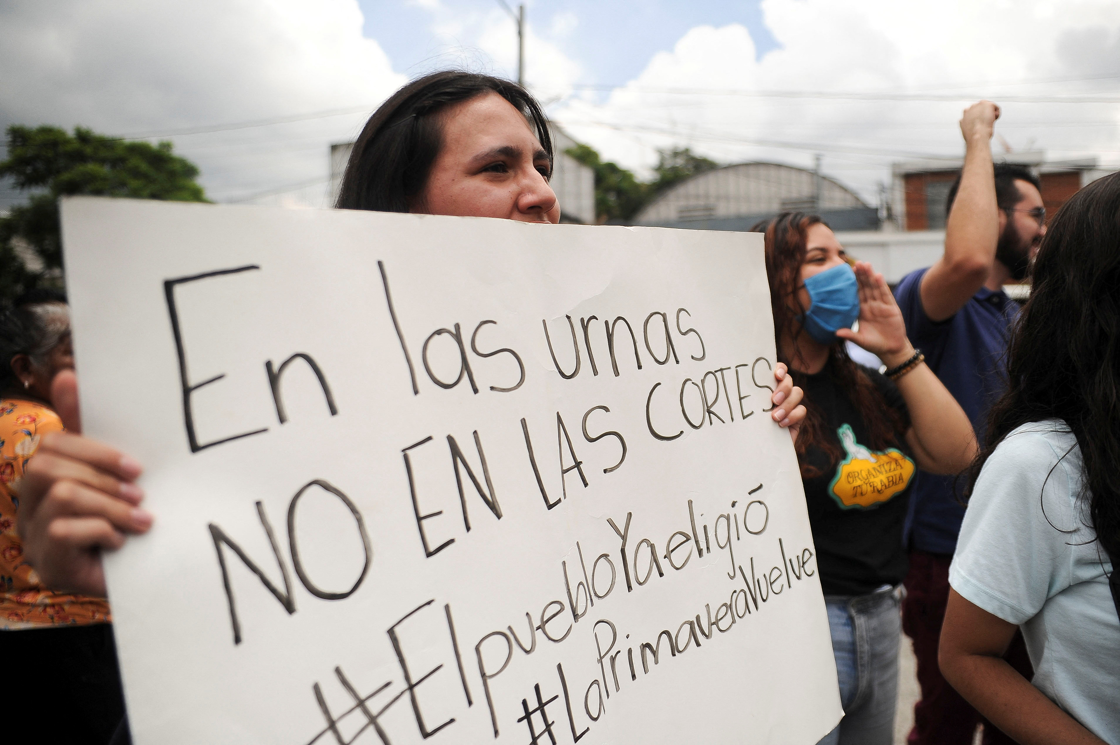 A woman holds a white poster with handwritten text at an outdoor rally.