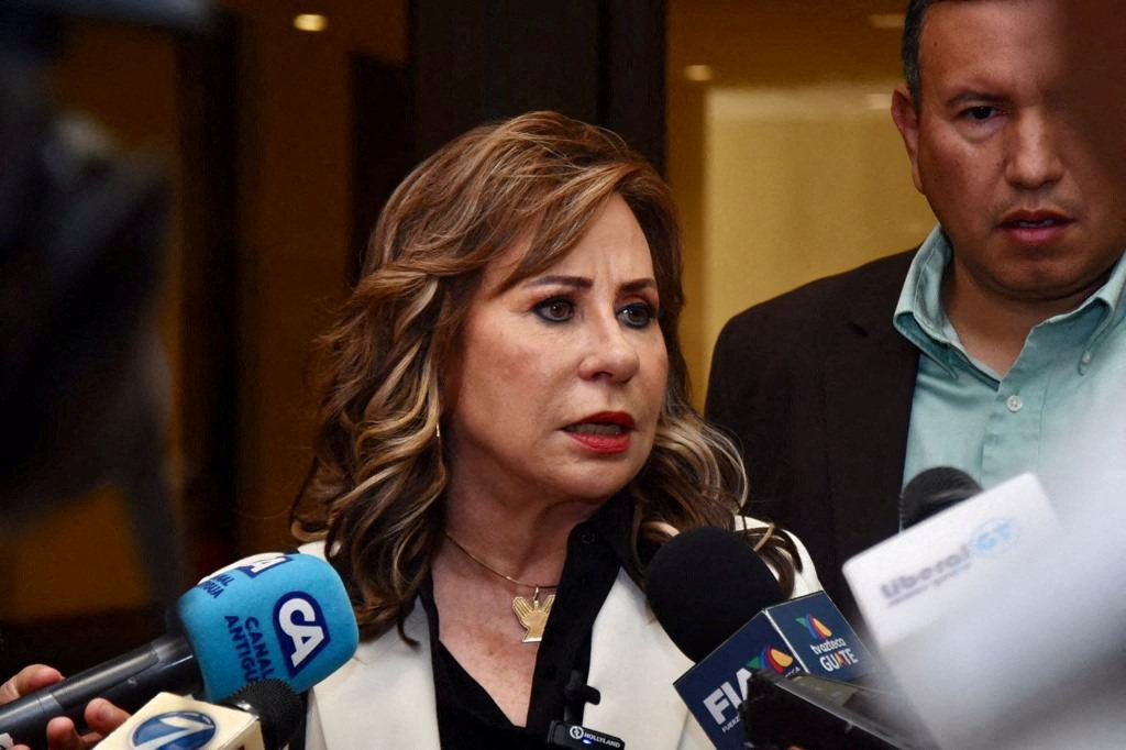 A woman in a white suit and black shirt speaks into a cluster of microphones.