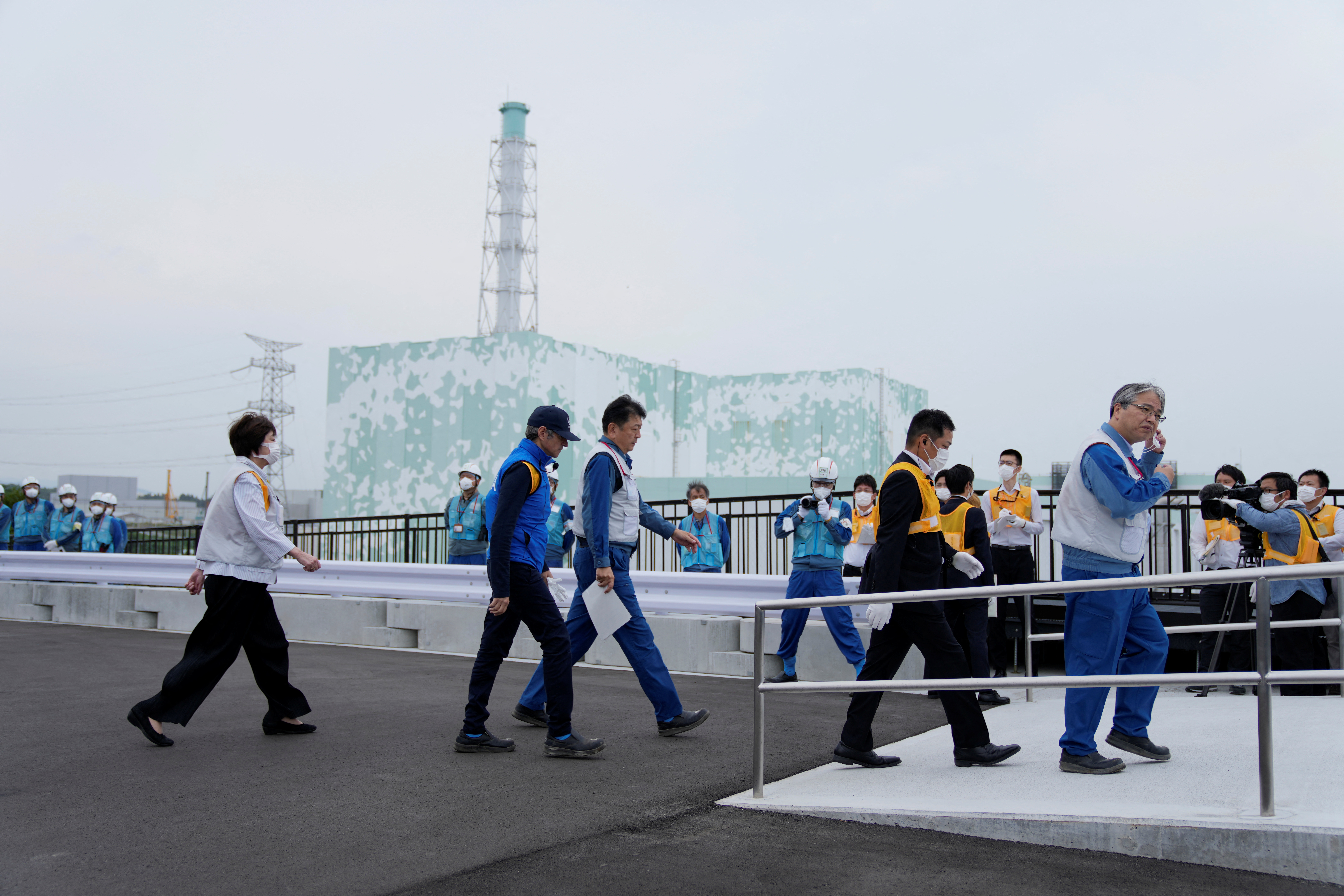 Rafael Grossi visiting the Fukushima power plant. The ruined building is in the distance behind him. He is walking and accompanied by power plant officials.