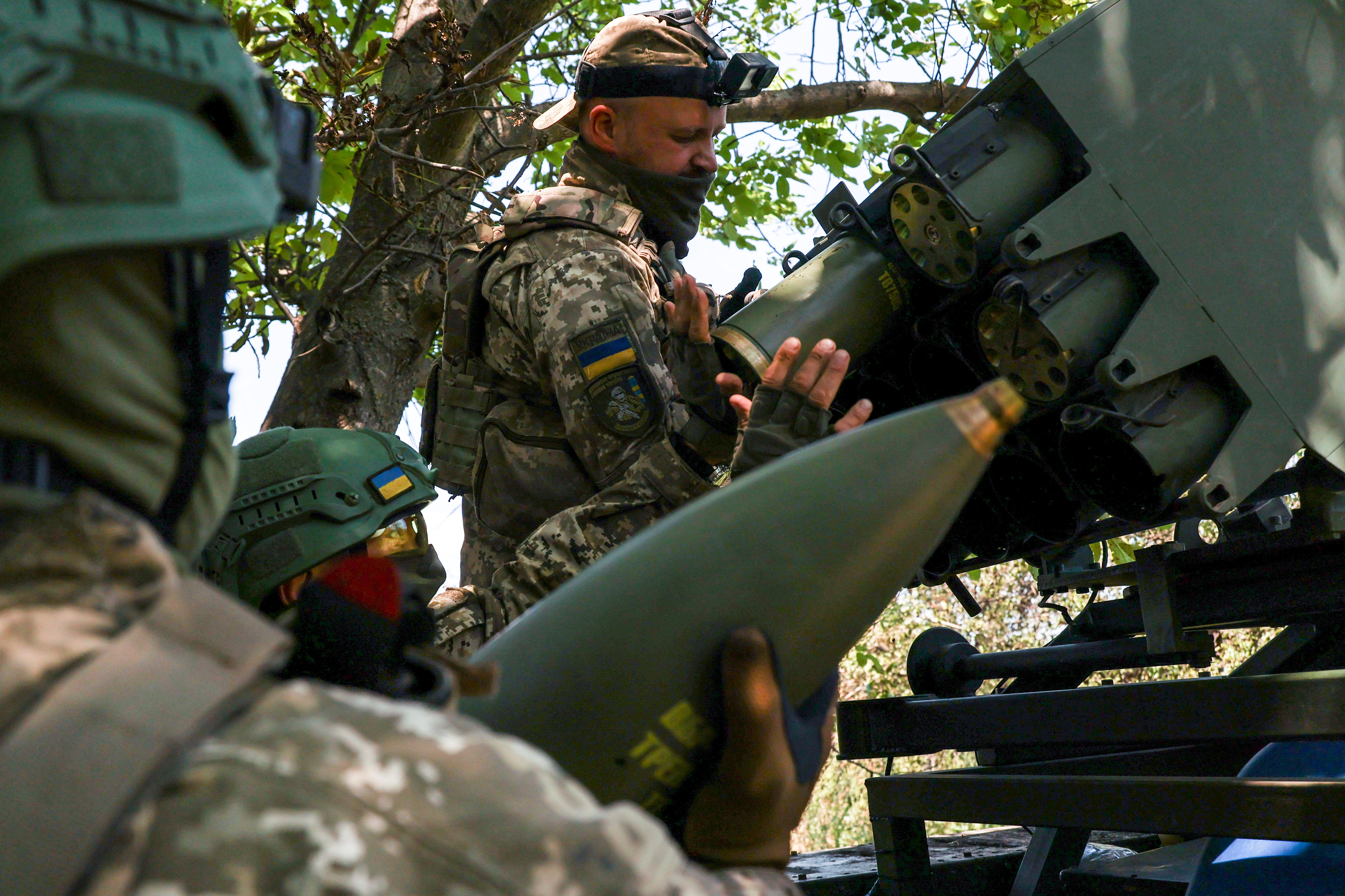 Three Ukrainian soldiers loading shells into a multiple launch rocket system. There are three of them. They are in a concealed position beneath trees.