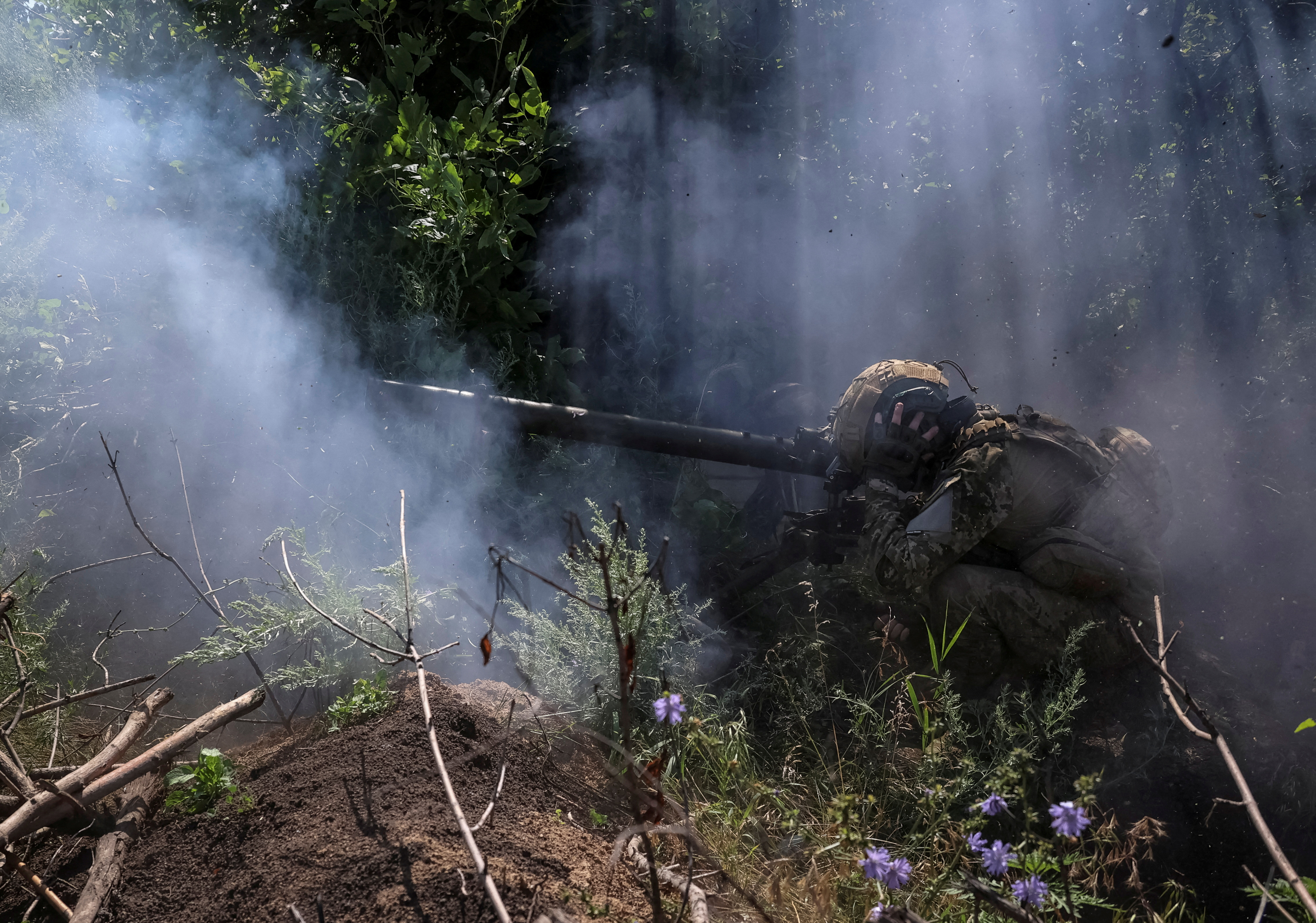 A Ukrainian soldier firing an anti-tank grenade launcher on the front line. He is crouched down, There are bushes around and some smoke.