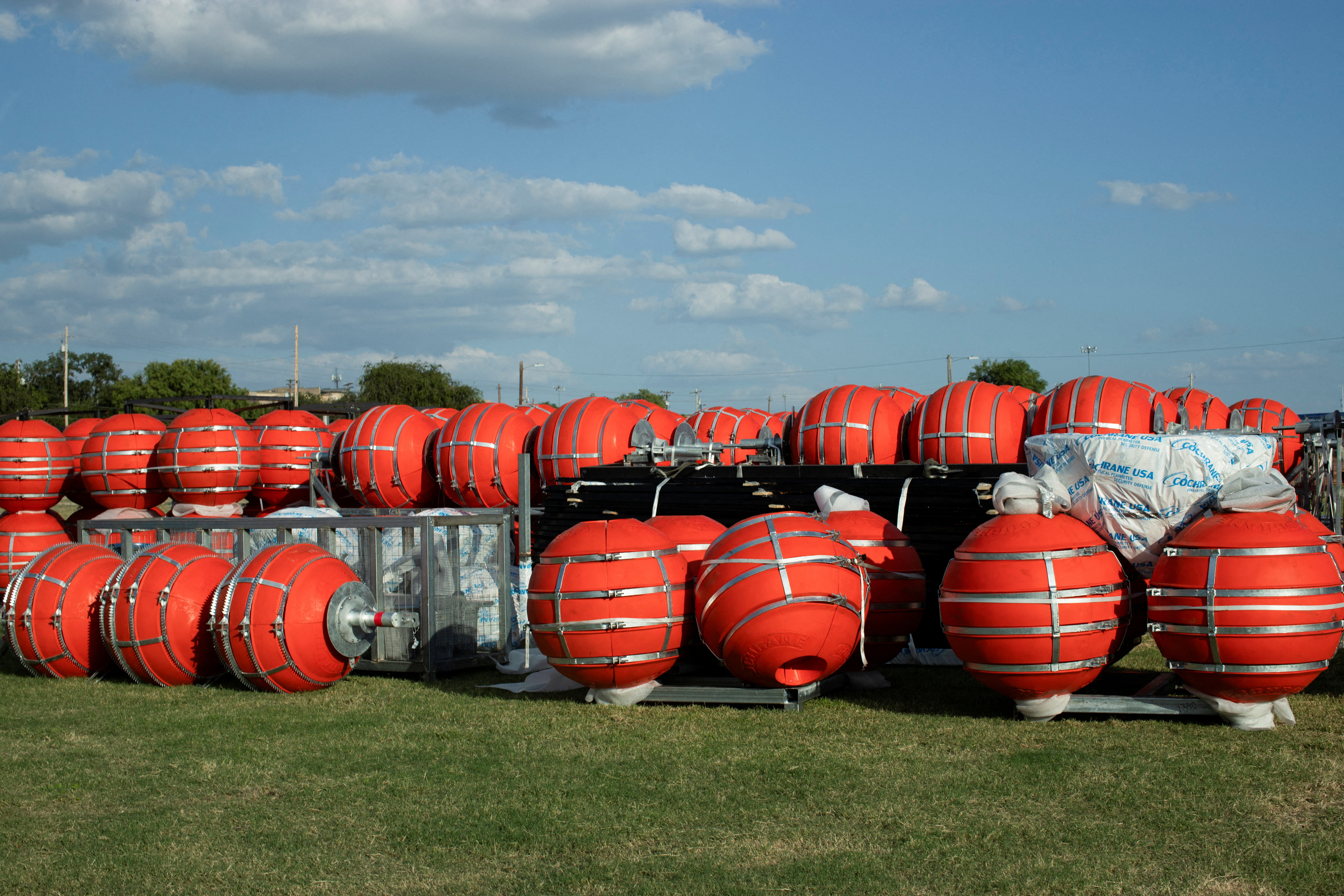 Giant orange buoys, wrapped with metal, stand stacked on a patch of grass.