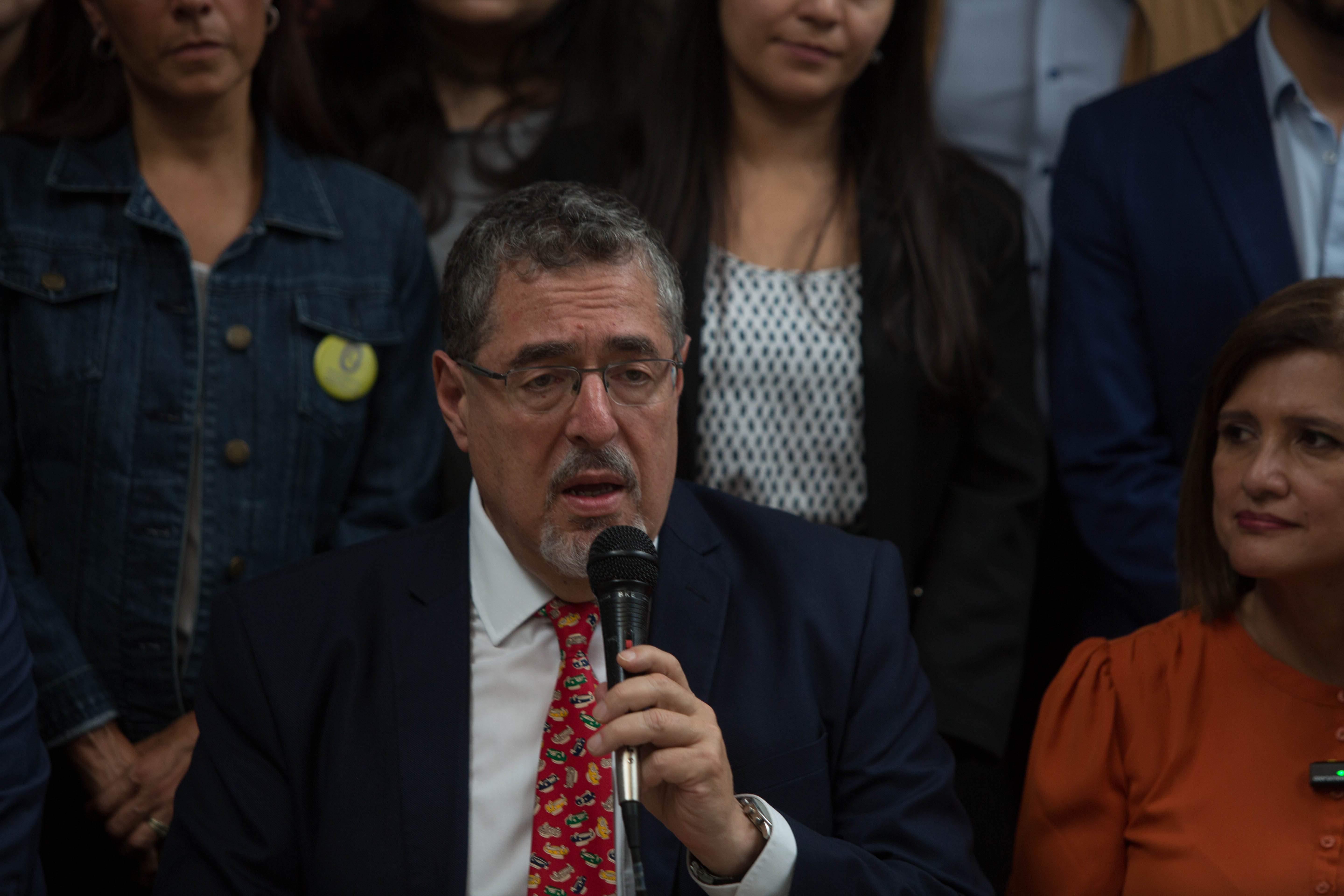 A man in a dark suit and red tie speaks into a microphone, surrounded by supporters