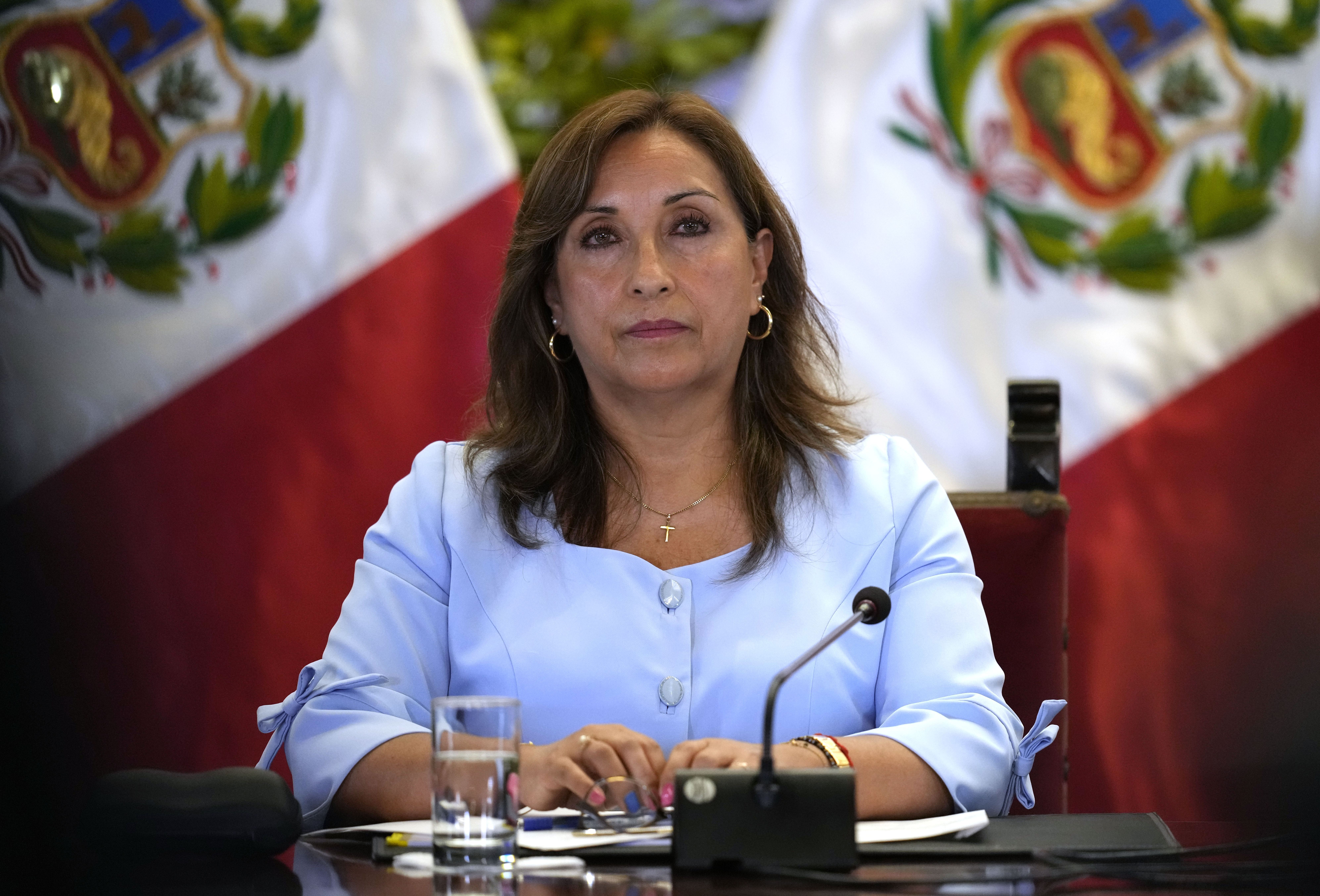 A woman sits at a table with a microphone in light blue dress clothes, surrounded by Peruvian flags.