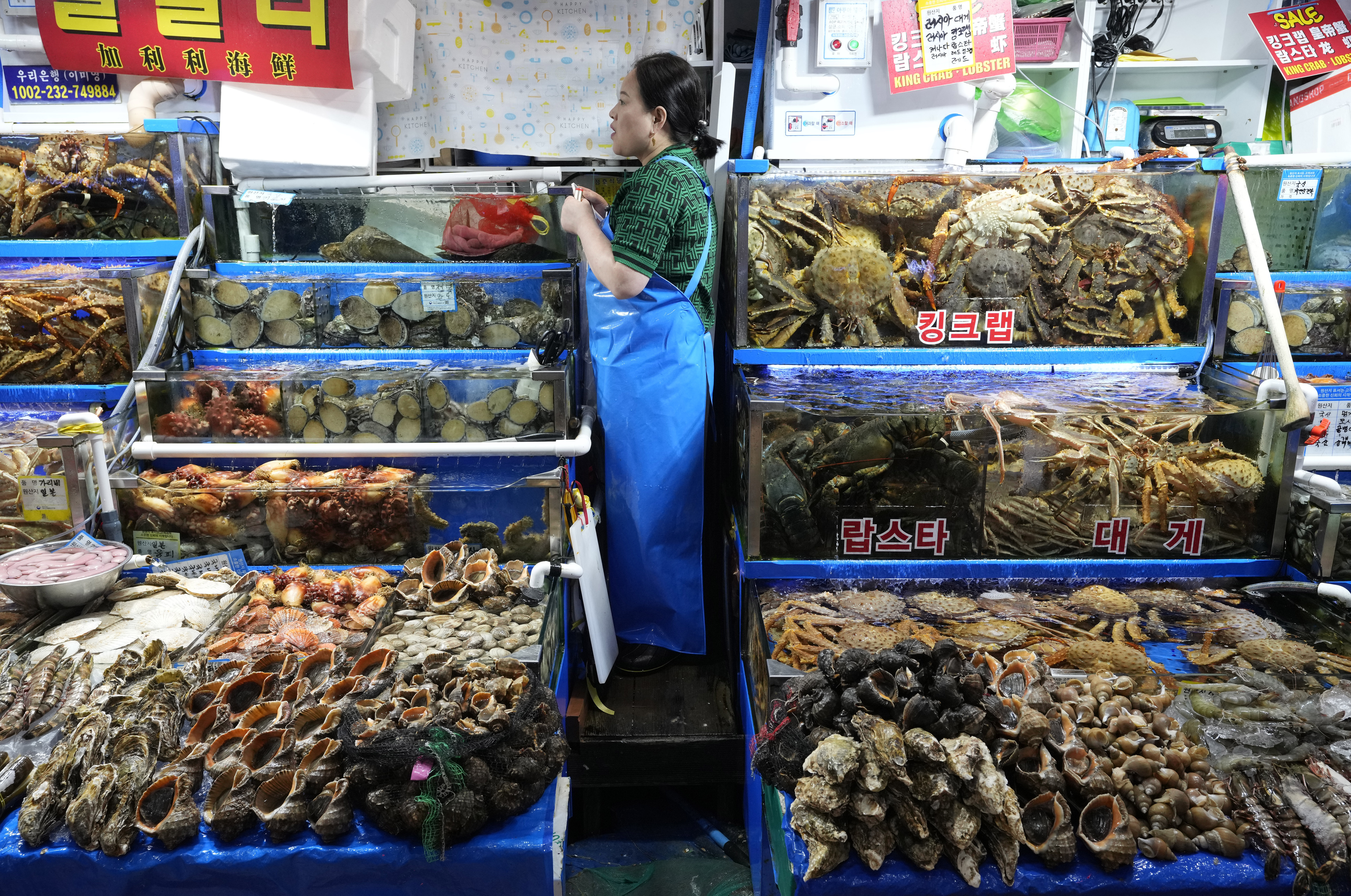 A woman standing between stalls laden with seafood in a Seoul market. She is wearing blue overalls.