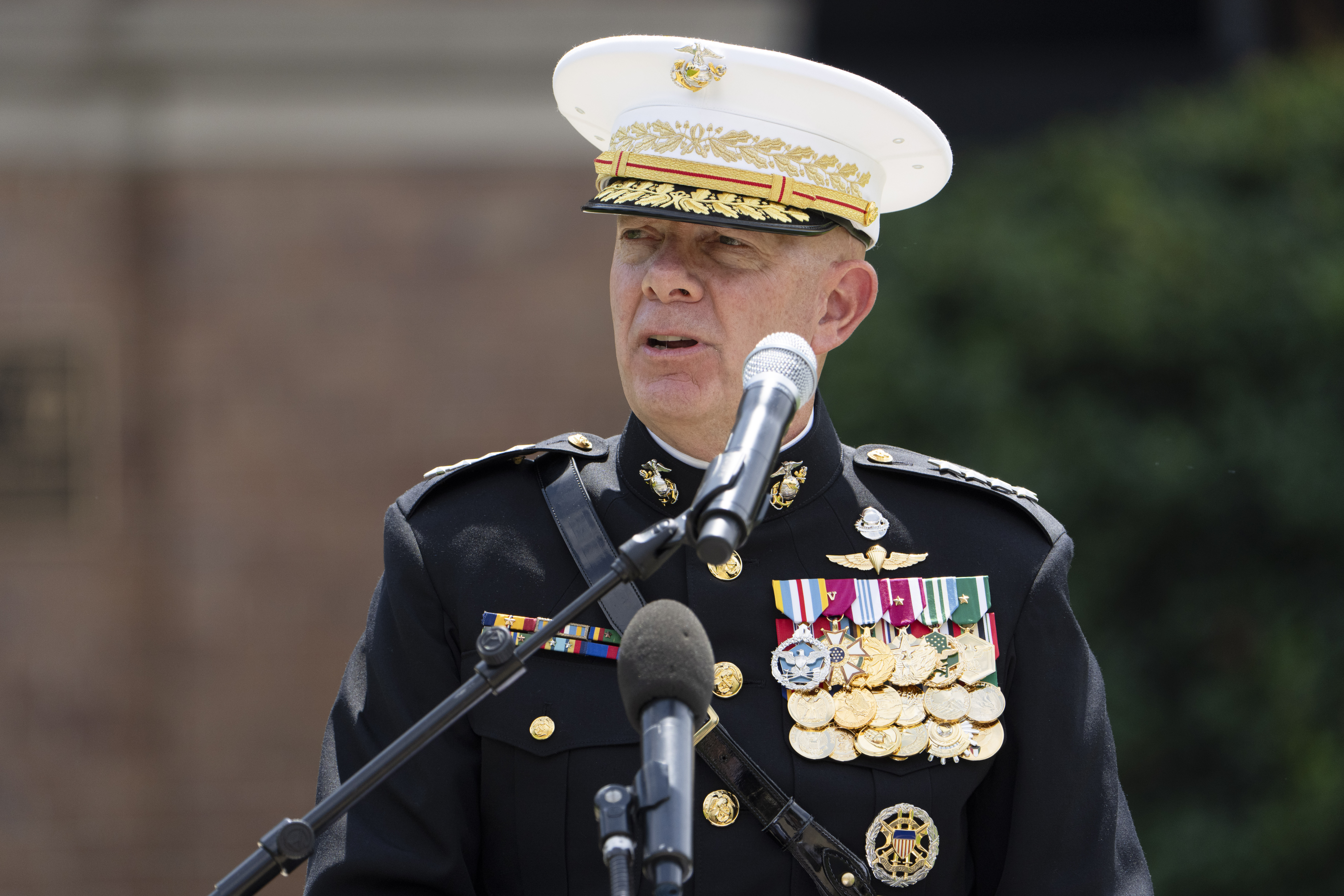 A man in a US Marine dress uniform — a white cap decorated with gold embroidered laurel leaves, and a black uniform studded with medals — gives a speech before a microphone.