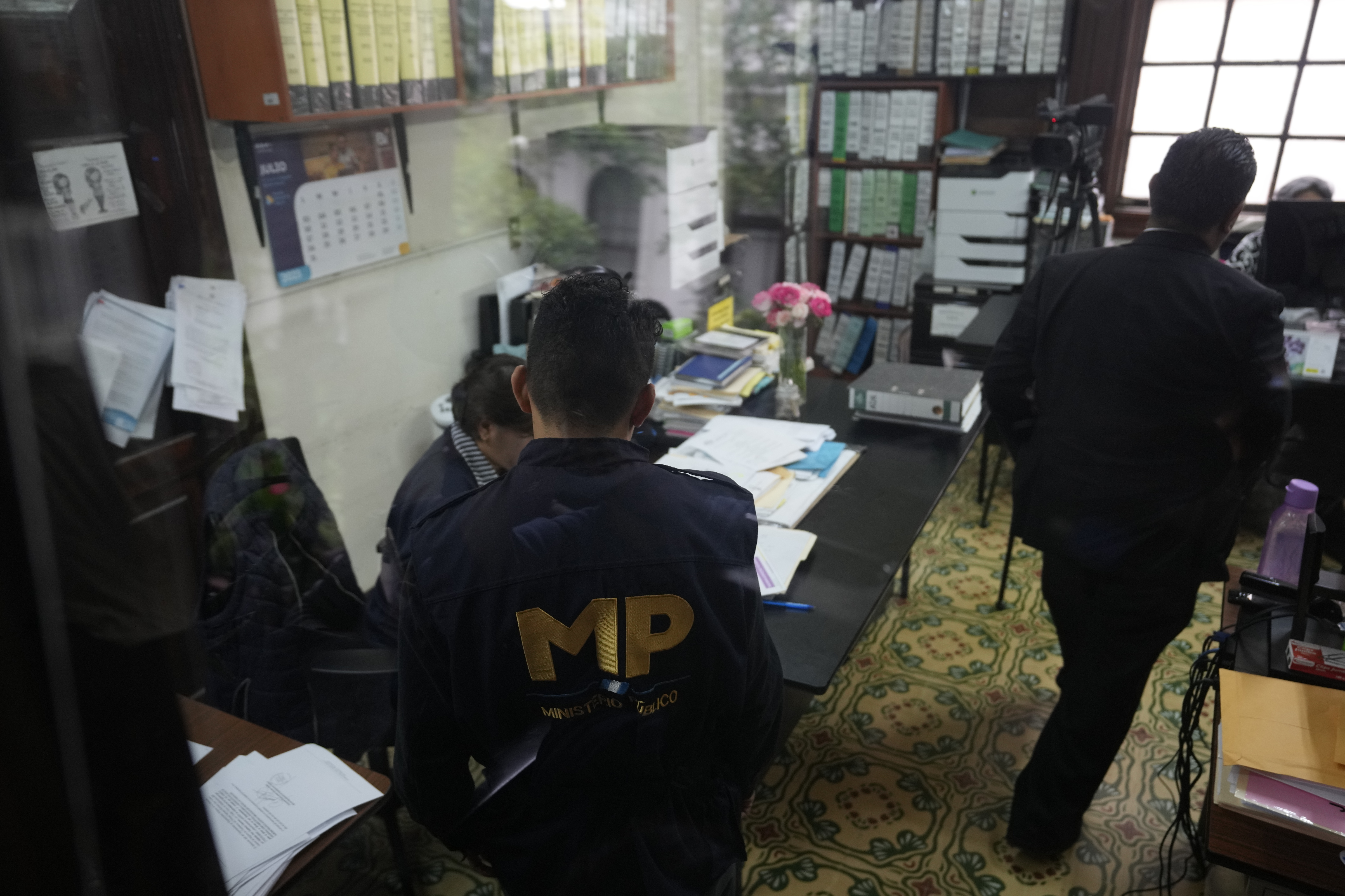 Men in black jackets labelled "MP" enter an office with files lining the walls.