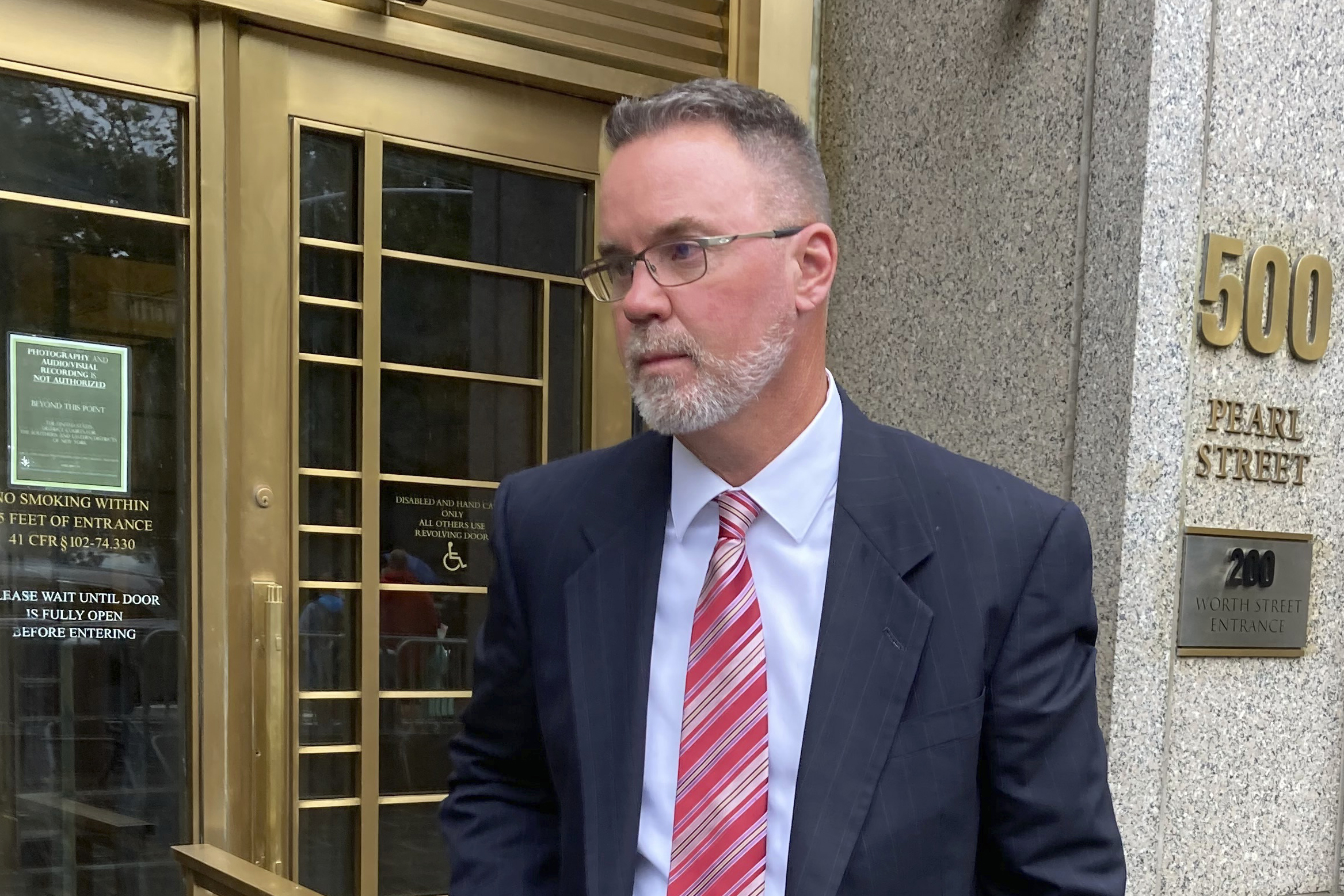 A man in a dark suit and red tie with glasses stands outside a courtroom door.