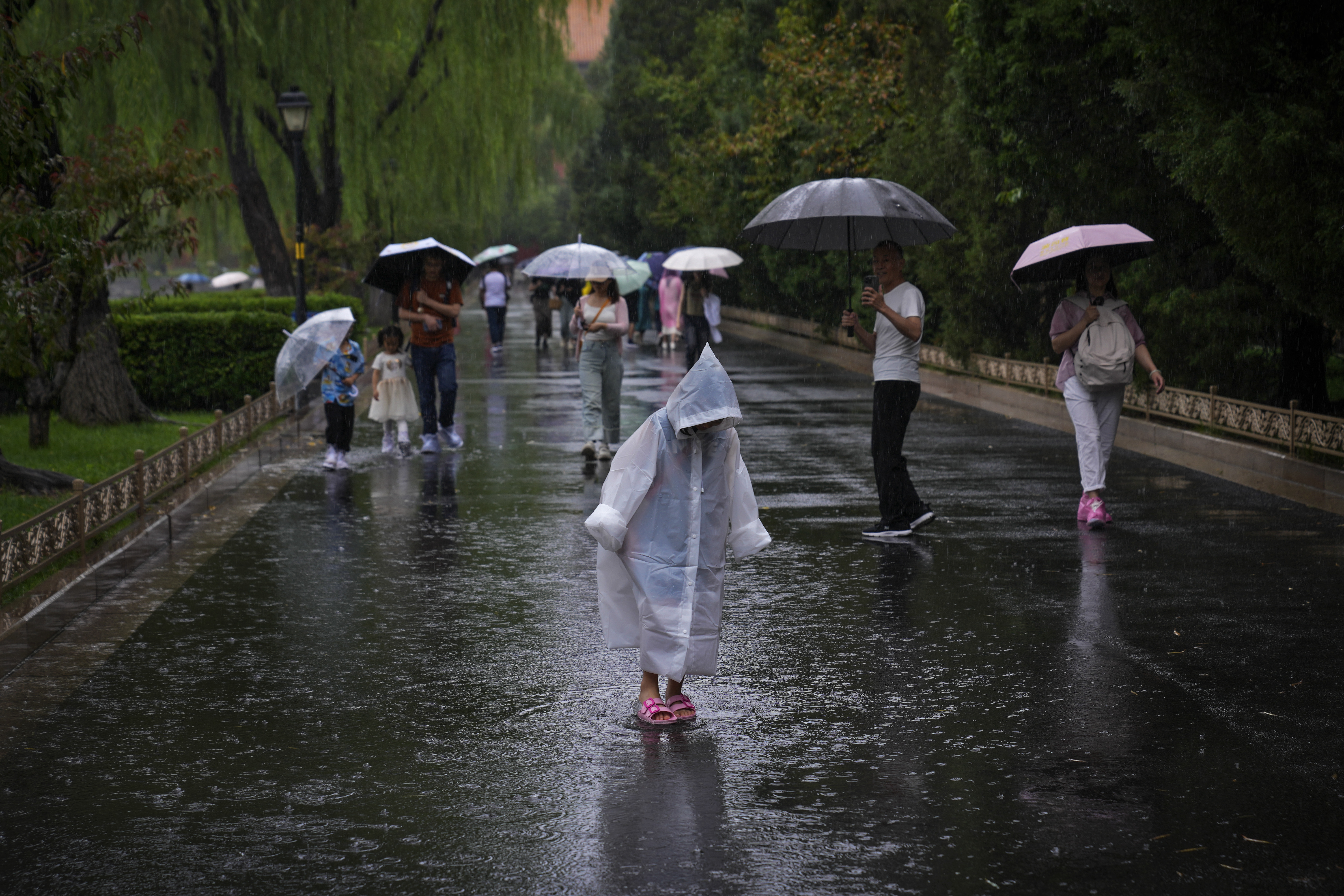 A child wearing a rain cover plays with rain water as visitors tour near the closed Forbidden City