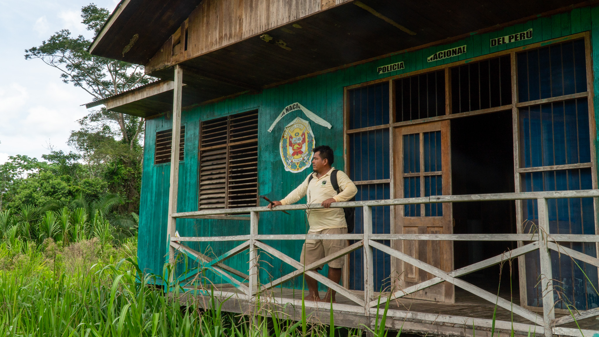 A man stands on the porch of a wooden building, with the seal of Peru painted on the side. The Amazon rainforest is visible in the background, and the building is surrounded by tall grass and shrubs.