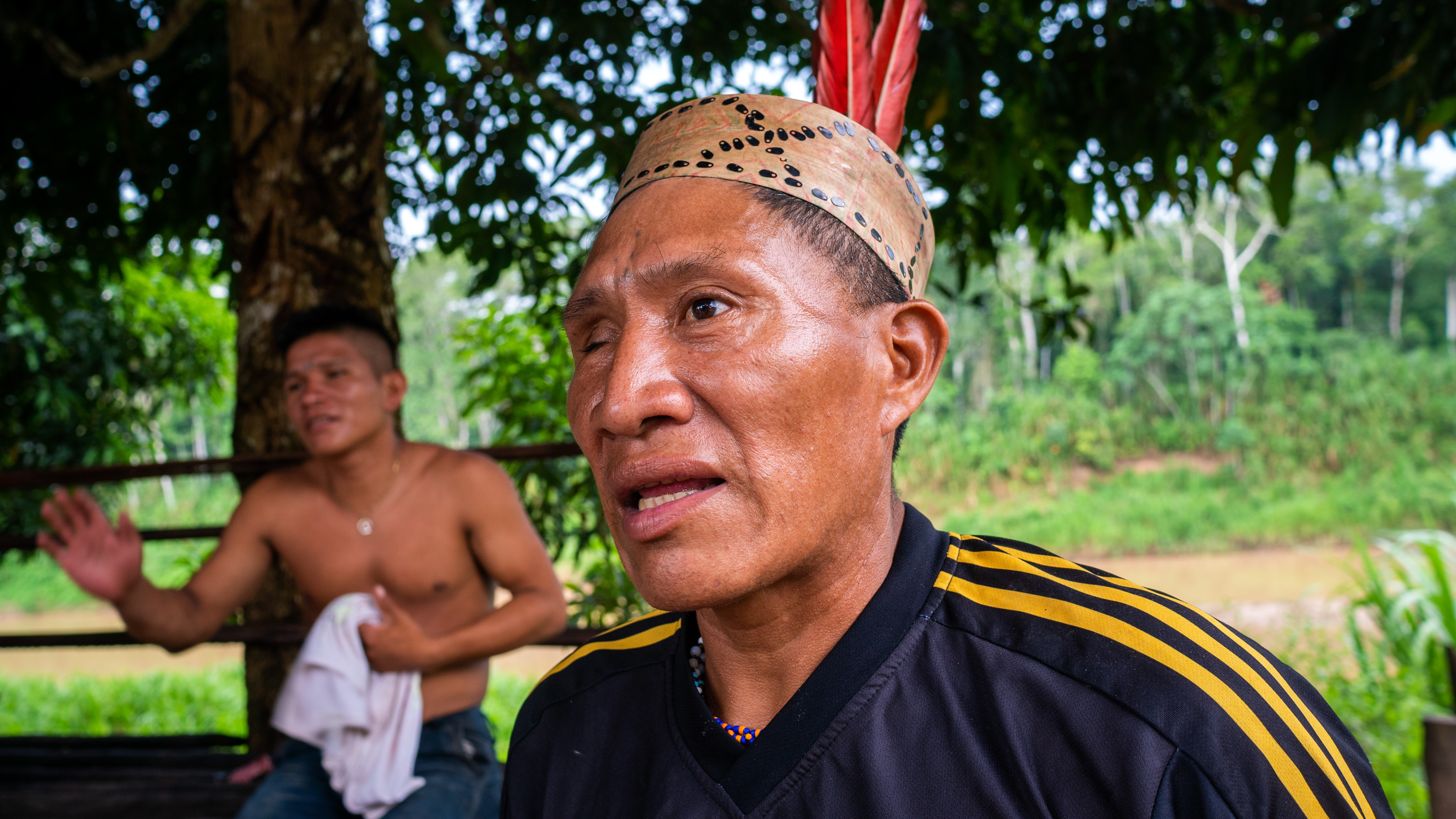 A man in a hat with red feathers and a shirt with stripes along the shoulders speaks to the camera. Behind him, a second, younger man is visible, having taken off his shirt. A forest is seen behind both of them.