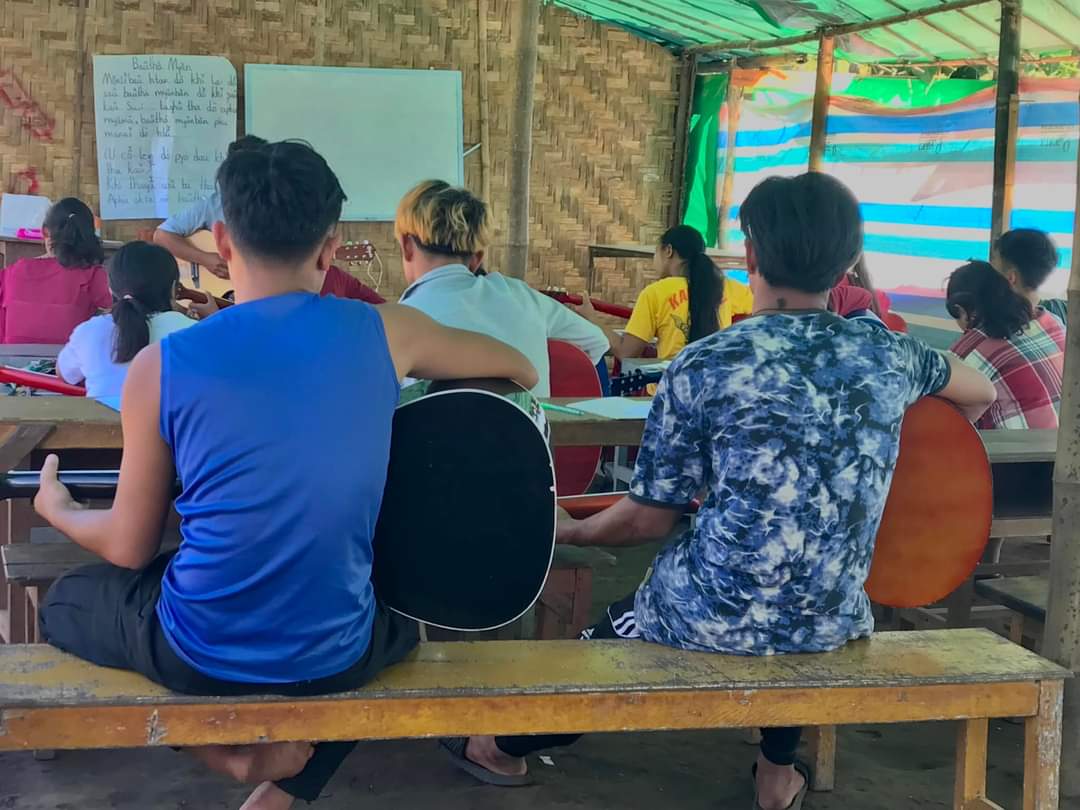Students learning the guitar in a community classroom. They are seated on benches/ Their backs are towards the camera.