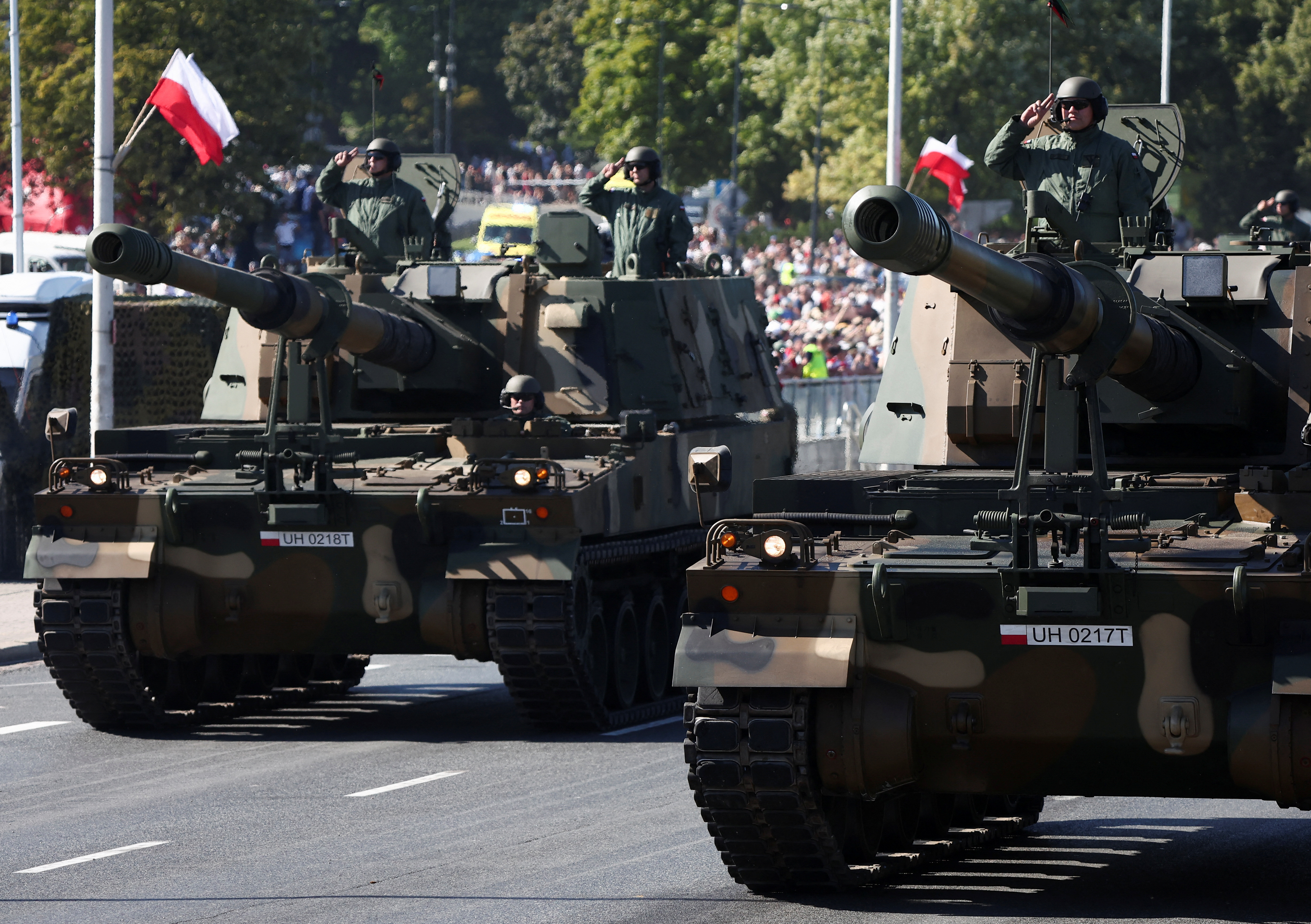 Tanks with armed soldiers standing inside and saluting roll through the streets. There are crowds behind them and Polish red and white flags are waving off poles