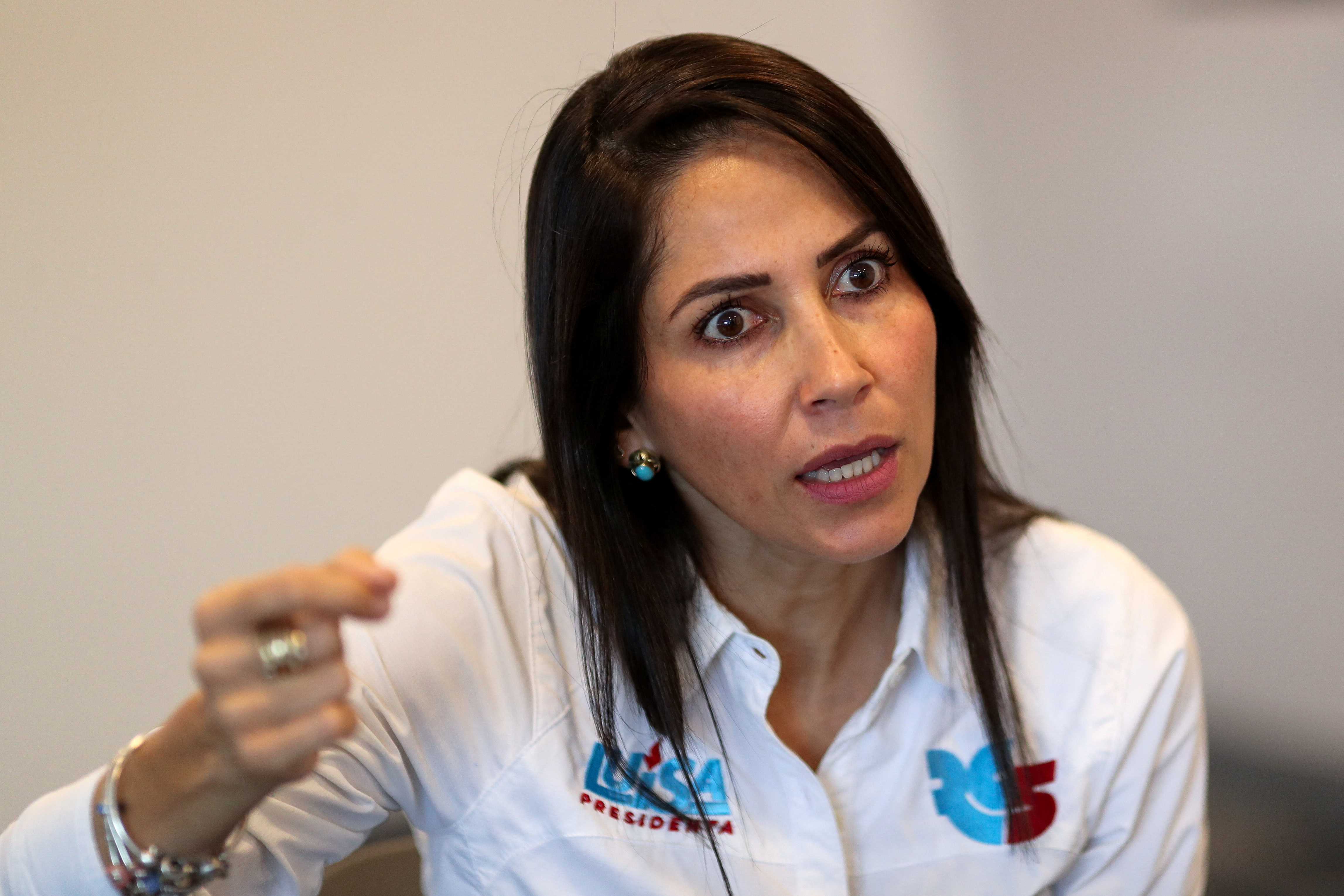 A woman in a white polo shirt sits in an empty room, speaking and gesturing passionately