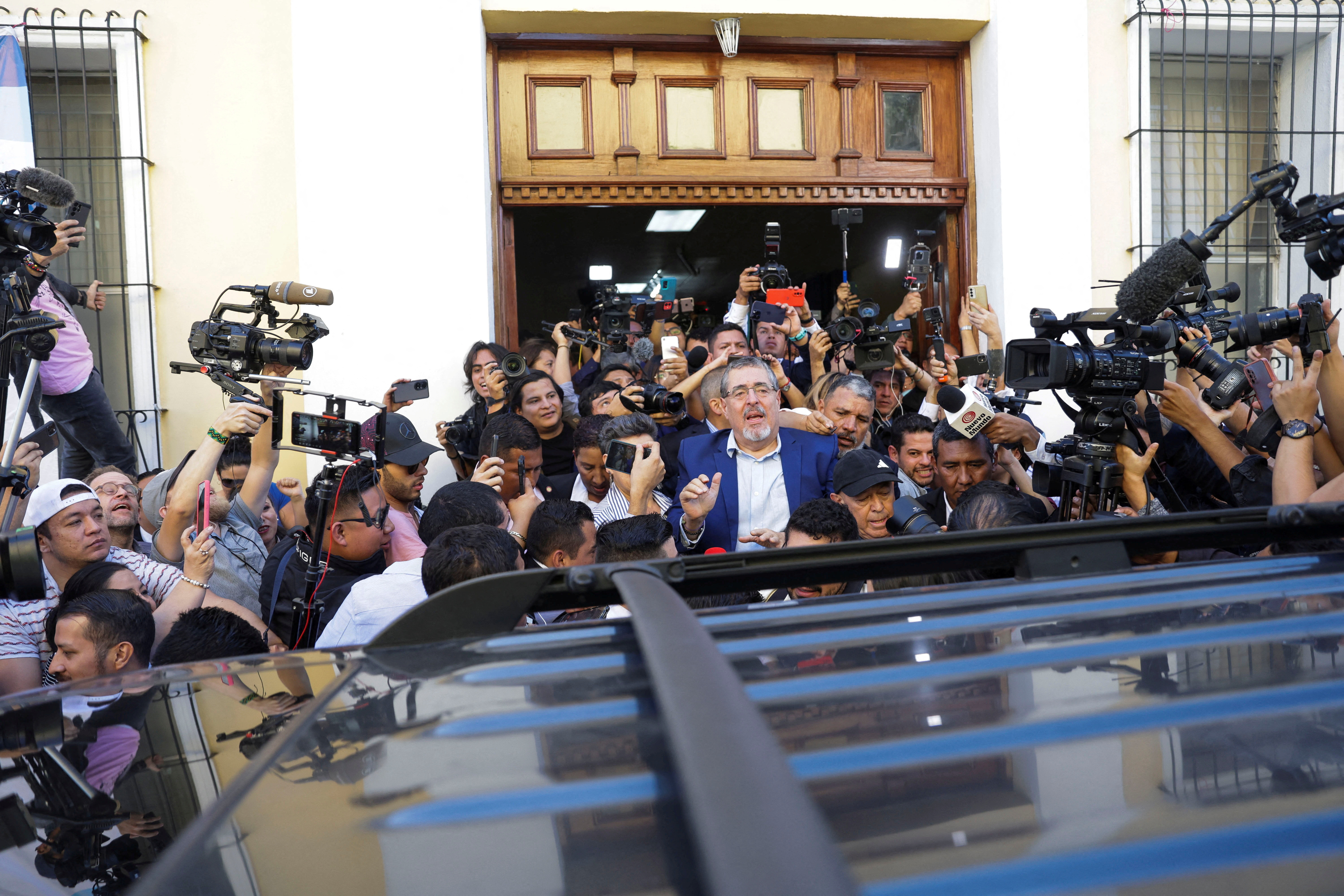 Bernardo Arevalo, dressed in a white collared shirt and a blue blazer, crosses a field of reporters to reach a waiting vehicle. Cameras are pointed at him from all directions.