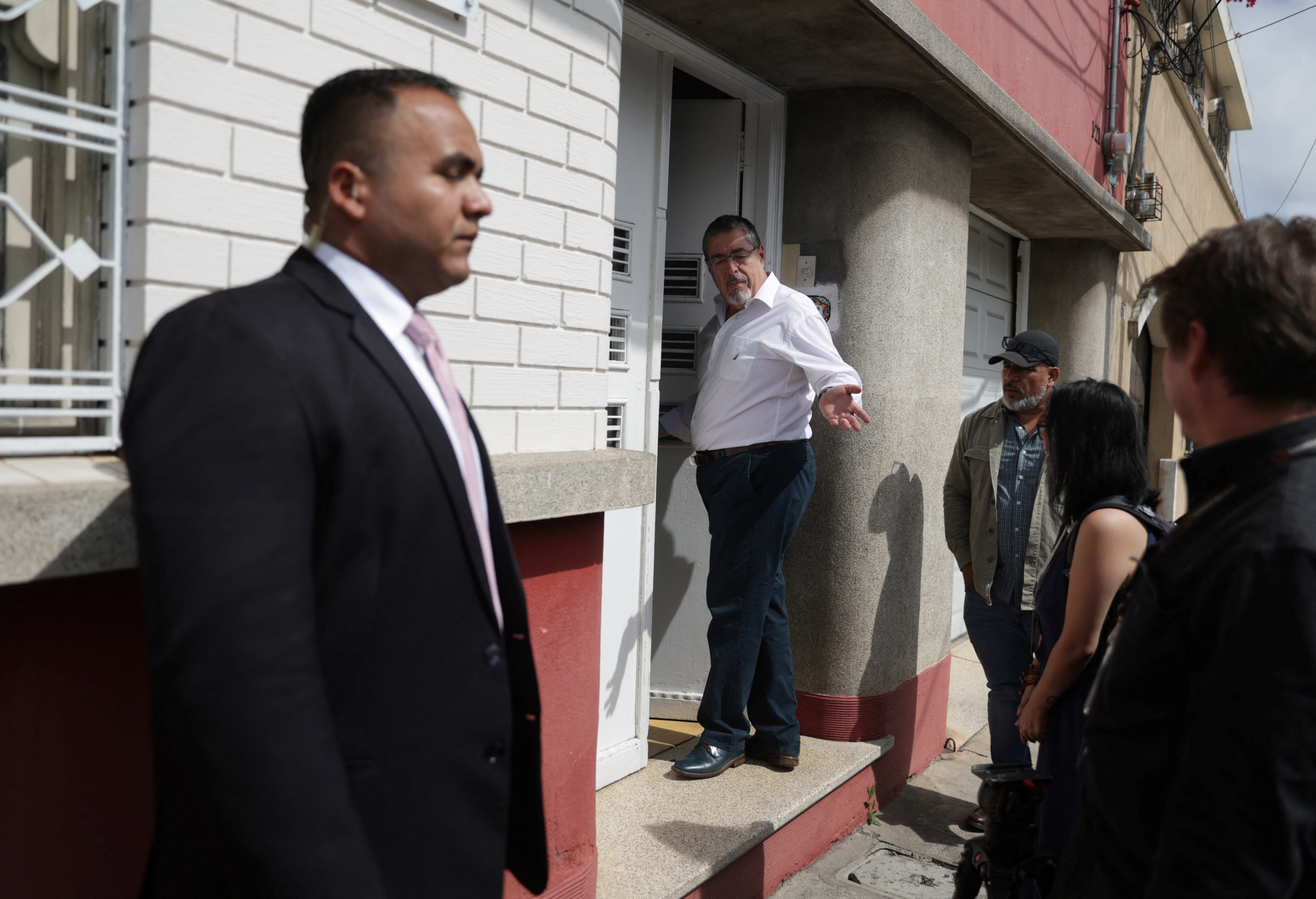 A security guard stands next to a white brick wall, as Bernardo Arevalo enters the doorway of his home, gesturing back to reporters on his doorstep.