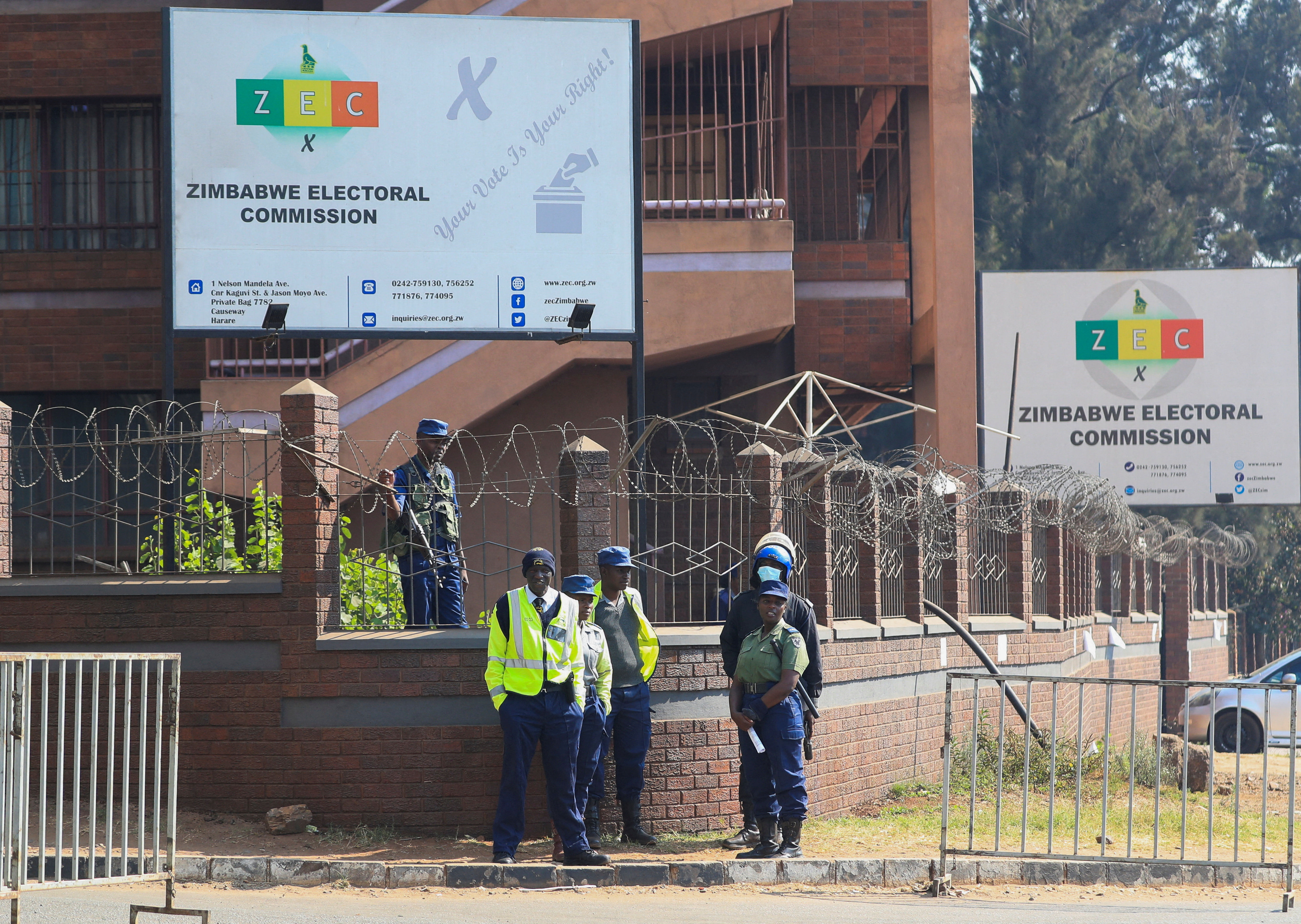 Police stand next to a barricade with razor wire, below a sign indicating the offices of the Zimbabwe Election Commission.