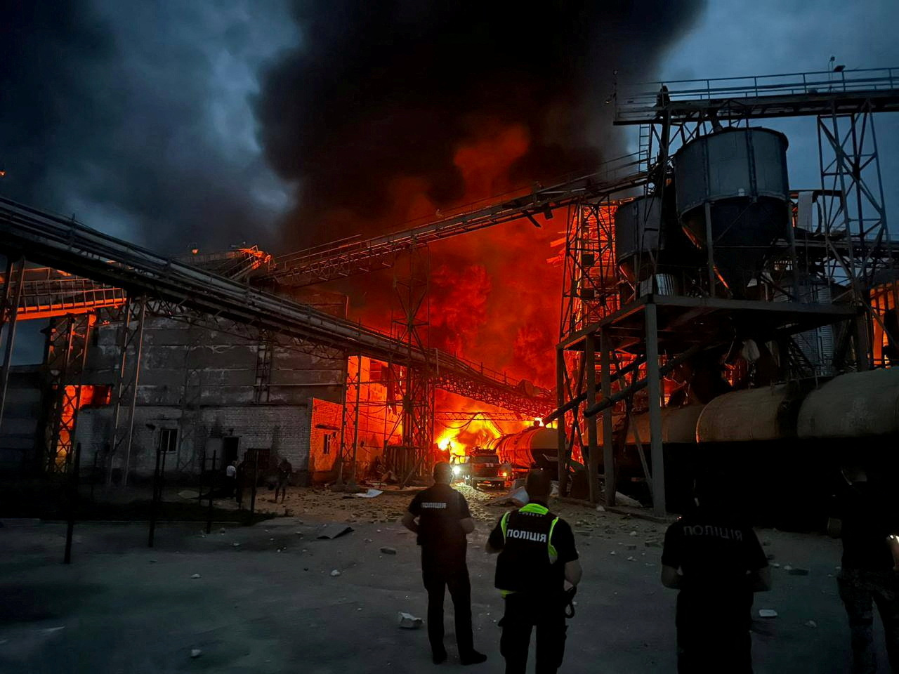 A vegetable oil factory on fire after a Russian missile attack. There are flames and black smoke. There are two rescue workers at the scene.