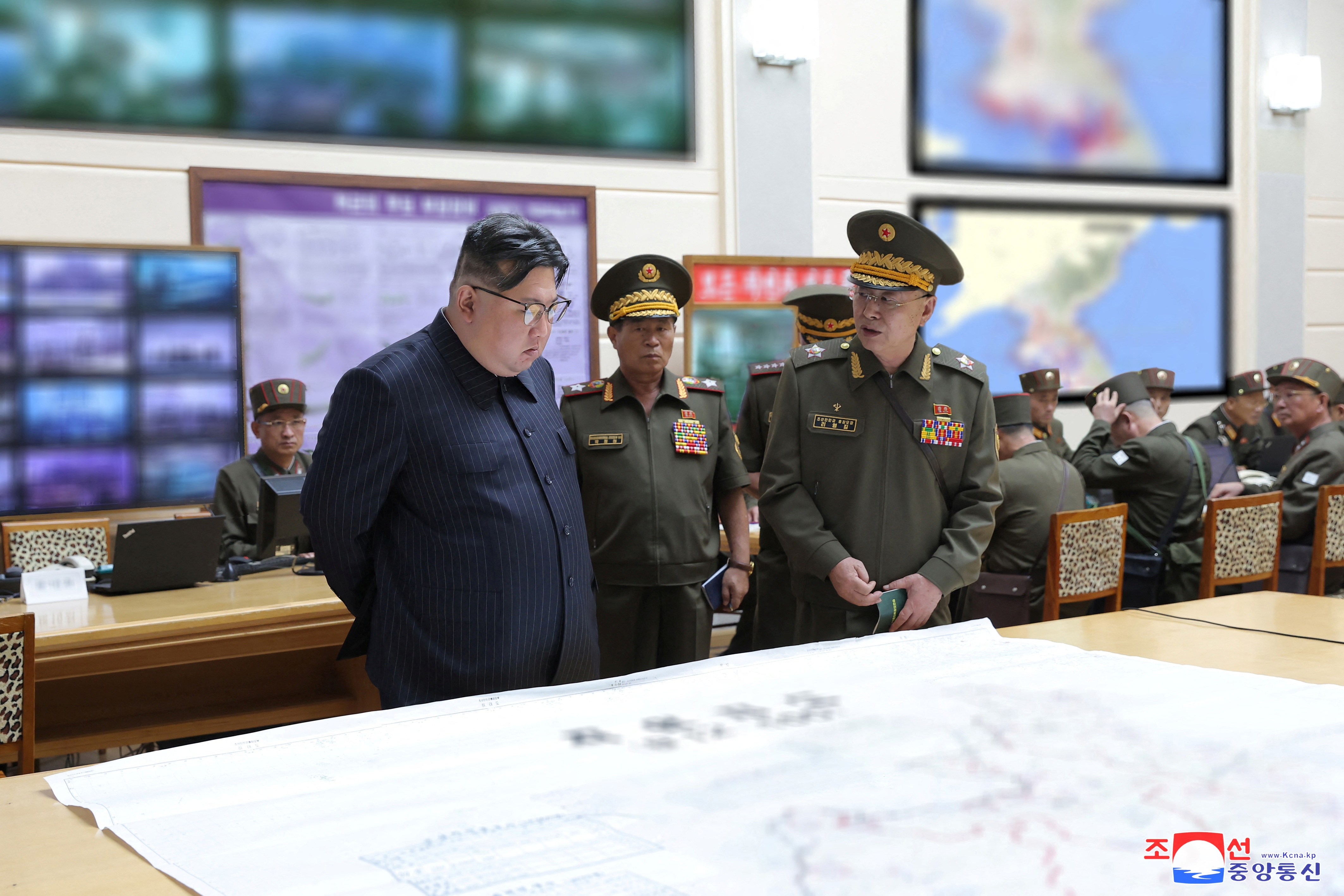 North Korean leader Kim Jong Un visiting the army training centre. He is looking at charts laid out on a table. Two officers are briefing him.