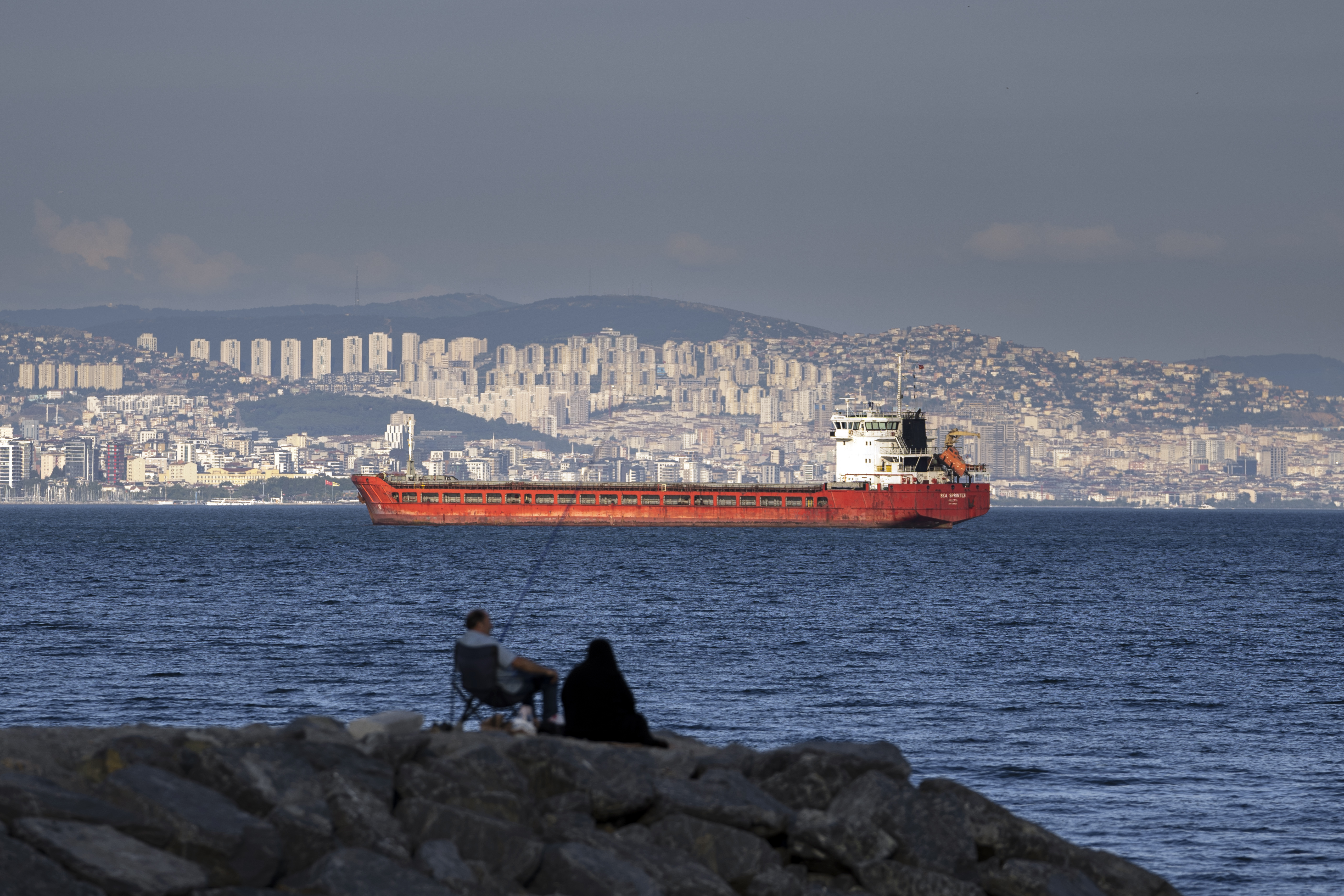 FILE - A family sit on a rock in front of a cargo ship anchors in the Marmara Sea awaits to access to cross the Bosphorus Straits in Istanbul, Turkey, on July 13, 2022. Shipping companies are not rushing to export millions of tons of trapped grain out of Ukraine, despite a breakthrough deal to provide safe corridors through the Black Sea. That is because the waters are mined, ship owners are still assessing the risks and many still have questions over how the deal will unfold. (AP Photo/Khalil Hamra, File)